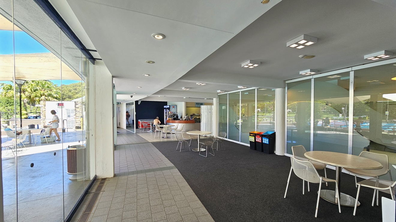 Movable tables and chairs are arranged to the right of the glass walls and in front of the cafe counter. The cafe features a timber and silver desk. An ice cream freezer and a fridge for cold drinks are positioned to the left and right of the counter. The floor is a combination of grey tiles and grey carpet. The ceiling is white with square and round fluorescent lights. Movable tables and chairs are arranged to the right of the glass walls and in front of the cafe counter. The cafe features a timber and silver desk. An ice cream freezer and a fridge for cold drinks are positioned to the left and right of the counter. The floor is a combination of grey tiles and grey carpet. The ceiling is white with square and round fluorescent lights.