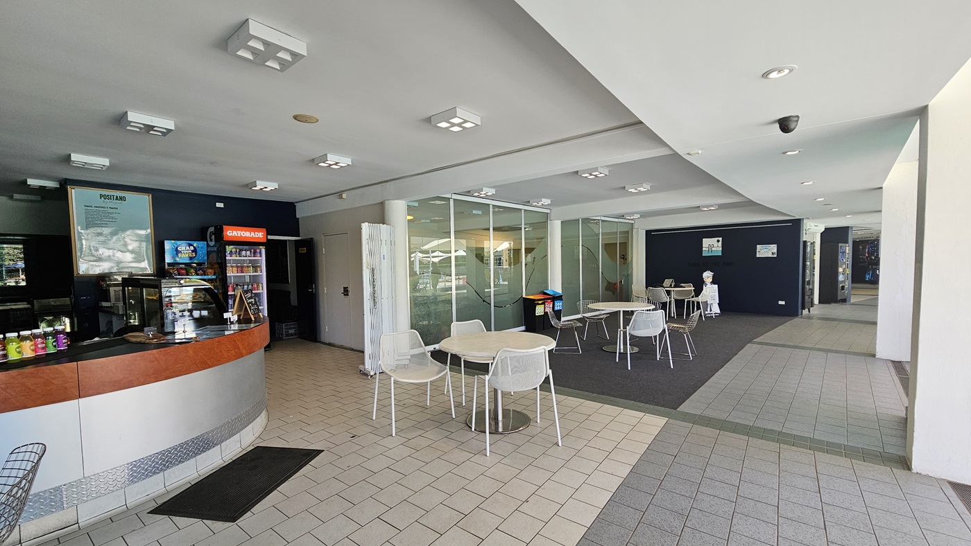Movable tables and chairs are arranged to the right of the glass walls and in front of the cafe counter. The cafe features a timber and silver desk. Food items and food racks are on top of the counter. A fridge displaying cold drinks stands on the right of the counter. The floor is a combination of grey tiles and grey carpet. The ceiling is white with square and round fluorescent lights. A black non-slip mat is also in place. Movable tables and chairs are arranged to the right of the glass walls and in front of the cafe counter. The cafe features a timber and silver desk. Food items and food racks are on top of the counter. A fridge displaying cold drinks stands on the right of the counter. The floor is a combination of grey tiles and grey carpet. The ceiling is white with square and round fluorescent lights. A black non-slip mat is also in place.