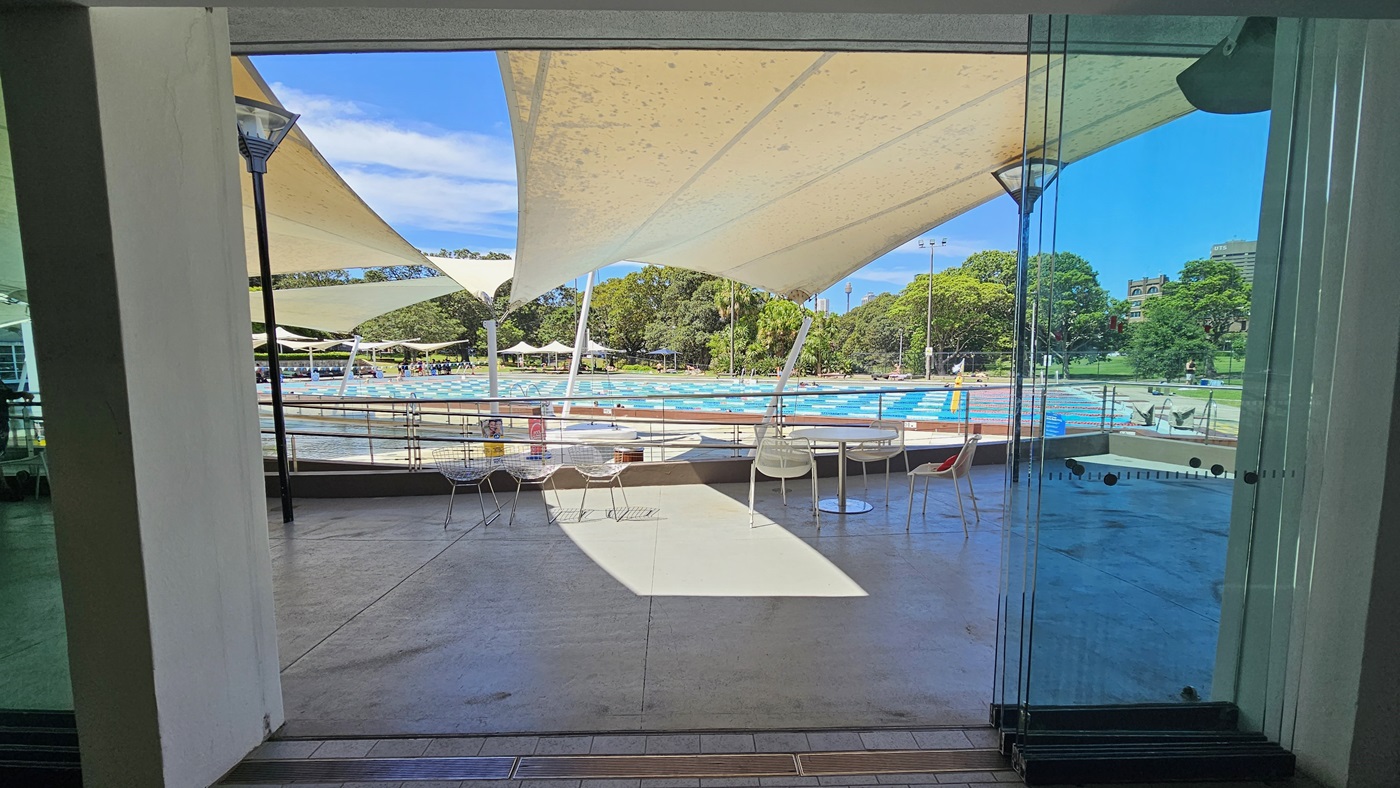 Movable tables and chairs are arranged in front of the cafe, near the pool deck. Multiple white cloth shades are placed overhead. Adjacent is the ramp entry equipped with handrails that leads to the pool. Lamp posts are also in place. Further behind is the 50m lap pool equipped with floating ropes. Umbrella shades and trees are visible further behind. Movable tables and chairs are arranged in front of the cafe, near the pool deck. Multiple white cloth shades are placed overhead. Adjacent is the ramp entry equipped with handrails that leads to the pool. Lamp posts are also in place. Further behind is the 50m lap pool equipped with floating ropes. Umbrella shades and trees are visible further behind.