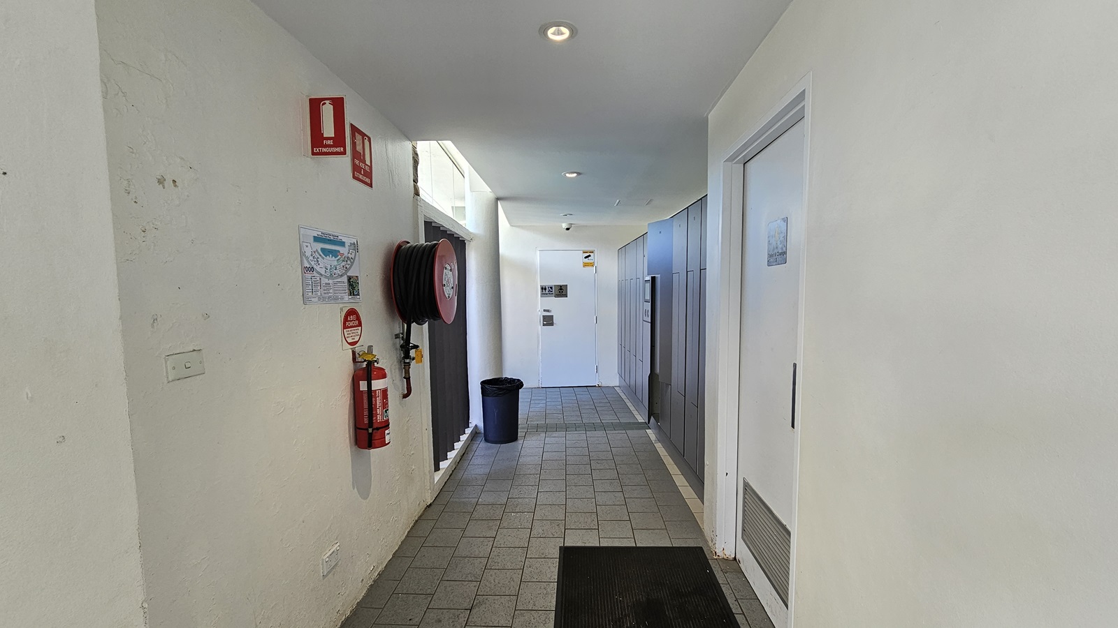 To the left is a fire hose reel and fire extinguisher mounted on the wall, equipped with signage. A poster bearing the site map is also affixed to the wall. To the right and further in are the doors leading to the accessible bathroom. A rubbish bin and a non-slip mat is also present. The floor has grey tiles, and the ceiling and wall are white with LED lights. To the left is a fire hose reel and fire extinguisher mounted on the wall, equipped with signage. A poster bearing the site map is also affixed to the wall. To the right and further in are the doors leading to the accessible bathroom. A rubbish bin and a non-slip mat is also present. The floor has grey tiles, and the ceiling and wall are white with LED lights.