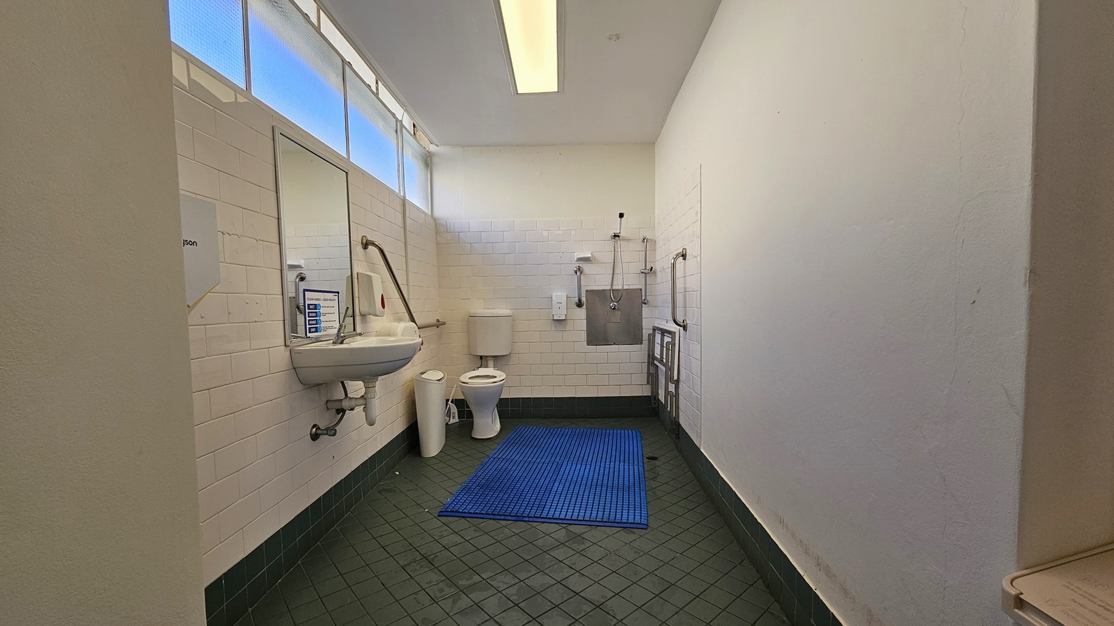 Inside the accessible bathroom, the toilet bowl equipped with handrails is in the left corner. Next to it is a rubbish bin. Adjacent to it is the shower, with hand rails and a foldable bench affixed to the right wall. To the left is a washbasin, mirrors, along with a liquid soap and tissue dispenser. The grey floor is equipped with a blue non-slippery mat, and the ceiling and wall are white with fluorescent lights. The upper left features a glass window. Inside the accessible bathroom, the toilet bowl equipped with handrails is in the left corner. Next to it is a rubbish bin. Adjacent to it is the shower, with hand rails and a foldable bench affixed to the right wall. To the left is a washbasin, mirrors, along with a liquid soap and tissue dispenser. The grey floor is equipped with a blue non-slippery mat, and the ceiling and wall are white with fluorescent lights. The upper left features a glass window.