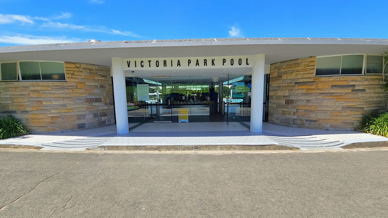 The facility entrance showcases a brown concrete facade with a portico displaying the facility's name in bold letters. Step-free access is available on both sides. Landscaped plants adorn the left and right areas. The entrance comprises a manual glass door with posters and a freestanding sign in the middle. The floor transitions from concrete to white bricks with grey accents. The facility entrance showcases a brown concrete facade with a portico displaying the facility's name in bold letters. Step-free access is available on both sides. Landscaped plants adorn the left and right areas. The entrance comprises a manual glass door with posters and a freestanding sign in the middle. The floor transitions from concrete to white bricks with grey accents.