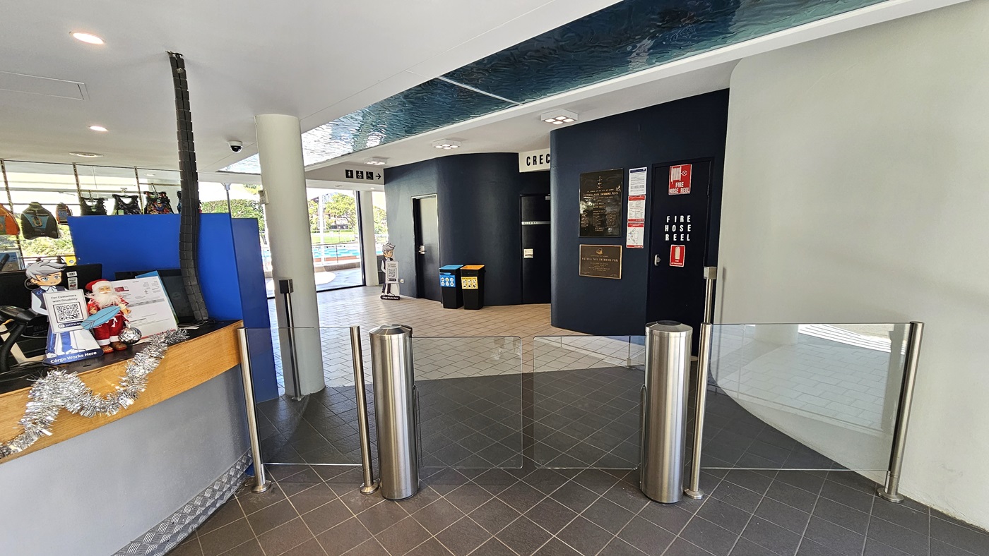 The reception desk, featuring a circular design with silver and timber elements, is centrally located. The counter is decorated with Christmas garland, card swipe machines, computers, and brochures. A turnstile glass gate is positioned to the right. Inside, a black wall displays posters and directional signage, with rubbish bins in place. The floor has grey tiles, and the ceiling is white with an aqua accent. Behind the desk, retail items are on display. The reception desk, featuring a circular design with silver and timber elements, is centrally located. The counter is decorated with Christmas garland, card swipe machines, computers, and brochures. A turnstile glass gate is positioned to the right. Inside, a black wall displays posters and directional signage, with rubbish bins in place. The floor has grey tiles, and the ceiling is white with an aqua accent. Behind the desk, retail items are on display.