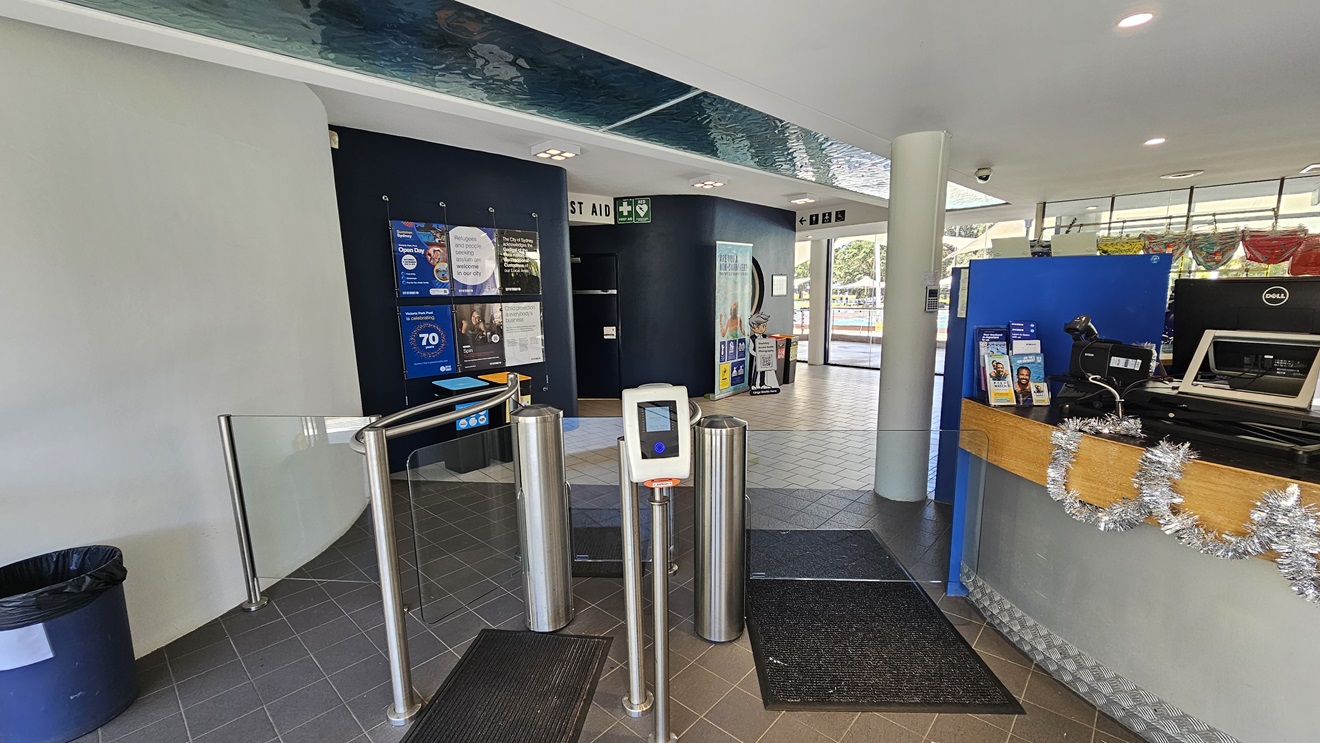 The centrally located reception desk has a circular design with silver and timber elements. The counter is adorned with Christmas garlands, computers, and brochures. To the left, a turnstile glass gate is equipped with a swiping machine and a non-slippery mat. Inside, a black wall displays posters and directional signage, with rubbish bins in place. The floor is covered in grey tiles, and the ceiling is white with an aqua accent. Behind the desk, retail items are on display. The centrally located reception desk has a circular design with silver and timber elements. The counter is adorned with Christmas garlands, computers, and brochures. To the left, a turnstile glass gate is equipped with a swiping machine and a non-slippery mat. Inside, a black wall displays posters and directional signage, with rubbish bins in place. The floor is covered in grey tiles, and the ceiling is white with an aqua accent. Behind the desk, retail items are on display.
