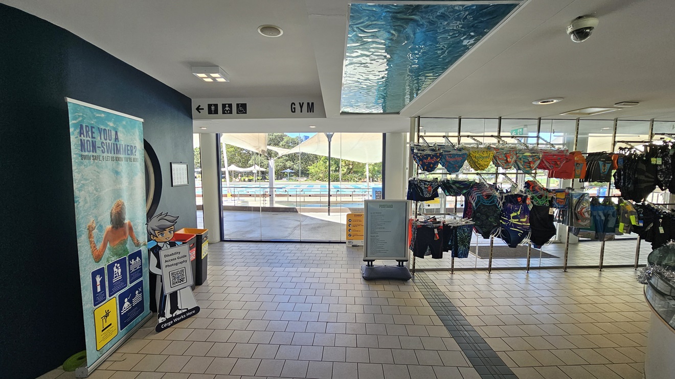 To the right is a retail area displaying swimwear. To the left, there's a large retractable banner, and two rubbish bins are positioned against the black wall. Directional signages are affixed to the upper left near the ceiling. Behind, a glass wall provides a view of the outdoor pool. The floor is covered in grey tiles, and the ceiling is white with an aqua accent. To the right is a retail area displaying swimwear. To the left, there's a large retractable banner, and two rubbish bins are positioned against the black wall. Directional signages are affixed to the upper left near the ceiling. Behind, a glass wall provides a view of the outdoor pool. The floor is covered in grey tiles, and the ceiling is white with an aqua accent.