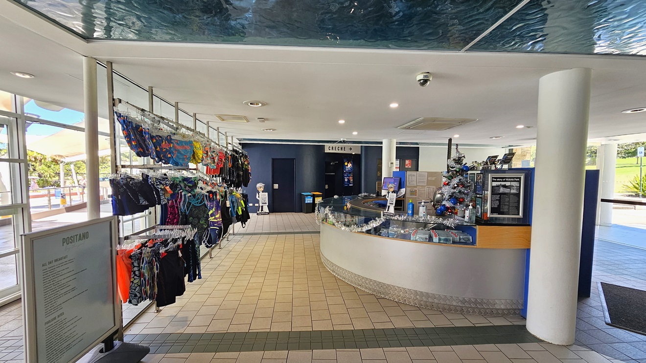 The reception desk, designed with a circular pattern using silver and timber elements, is located on the right. The counter is adorned with Christmas garland, computers, and brochures. On the left, swimwear is displayed for sale, along with a banner for the Positano cafe. The floor has brown tiles, and the ceiling is white with an aqua accent. The reception desk, designed with a circular pattern using silver and timber elements, is located on the right. The counter is adorned with Christmas garland, computers, and brochures. On the left, swimwear is displayed for sale, along with a banner for the Positano cafe. The floor has brown tiles, and the ceiling is white with an aqua accent.
