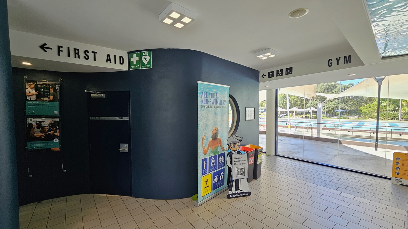 To the left, there's a black wall with posters and directional signages. A "First Aid" sign is affixed to the upper left. A large retractable banner is against the black wall with rubbish bins beside it. To the right is a glass wall and a yellow sign. Behind the glass wall is the outdoor aquatic facility. The floor is covered in grey tiles, and the ceiling is white with an aqua accent. To the left, there's a black wall with posters and directional signages. A "First Aid" sign is affixed to the upper left. A large retractable banner is against the black wall with rubbish bins beside it. To the right is a glass wall and a yellow sign. Behind the glass wall is the outdoor aquatic facility. The floor is covered in grey tiles, and the ceiling is white with an aqua accent.