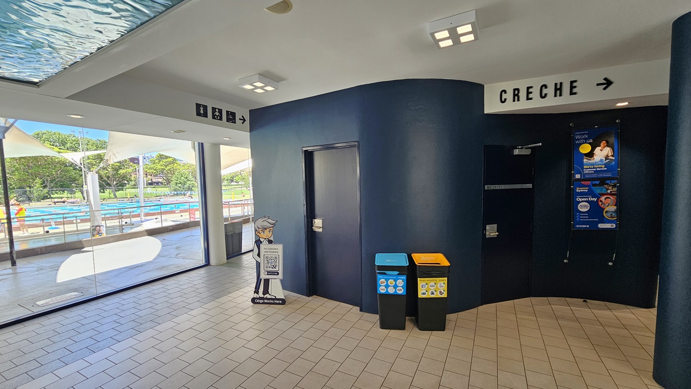 To the right, there's a black wall with posters and directional signages. A "Creche" sign is affixed to the upper right, and accessible bathroom signs are affixed to the upper left. Two rubbish bins are placed against the black wall. To the left is a glass wall, behind which is the outdoor aquatic facility. The floor is covered in grey tiles, and the ceiling is white with an aqua accent. To the right, there's a black wall with posters and directional signages. A "Creche" sign is affixed to the upper right, and accessible bathroom signs are affixed to the upper left. Two rubbish bins are placed against the black wall. To the left is a glass wall, behind which is the outdoor aquatic facility. The floor is covered in grey tiles, and the ceiling is white with an aqua accent.