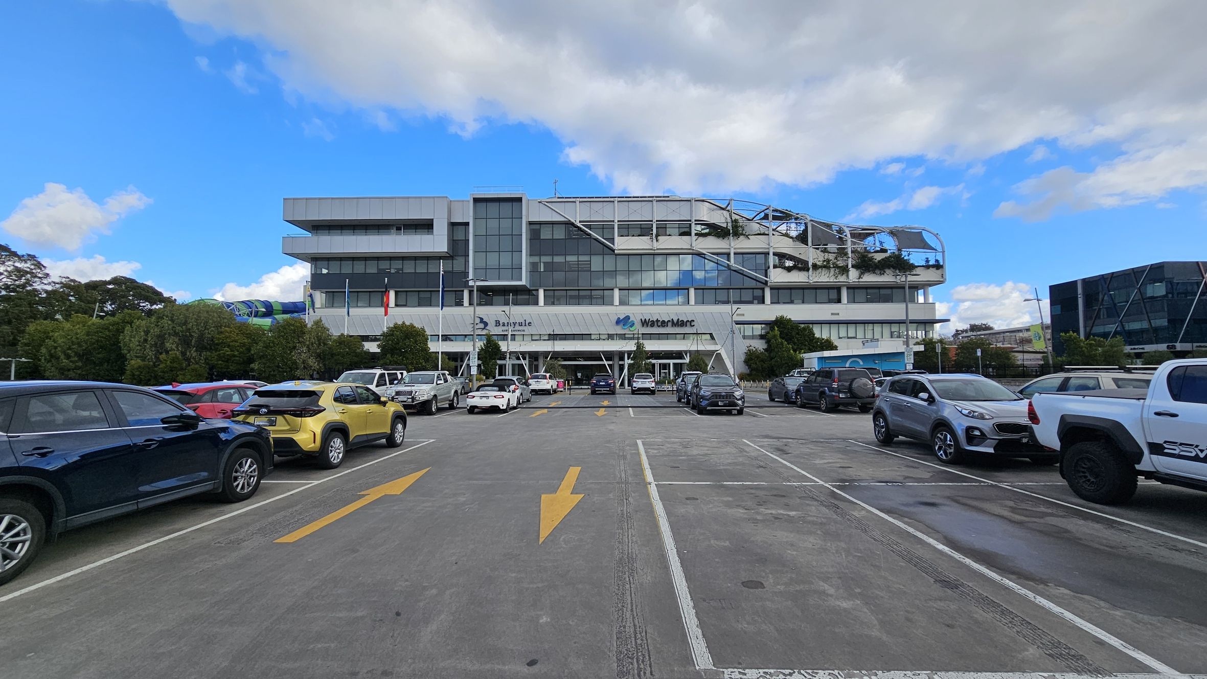 A large, modern building with a glass facade is in the centre, featuring the names "Banyule" and "WaterMarc" on the front. The foreground shows a car park with various vehicles, including a yellow car and a white truck. Yellow directional arrows are painted on the concrete surface. Trees and bushes line the perimeter of the car park, and another building with a geometric design is visible on the right. A large, modern building with a glass facade is in the centre, featuring the names "Banyule" and "WaterMarc" on the front. The foreground shows a car park with various vehicles, including a yellow car and a white truck. Yellow directional arrows are painted on the concrete surface. Trees and bushes line the perimeter of the car park, and another building with a geometric design is visible on the right.