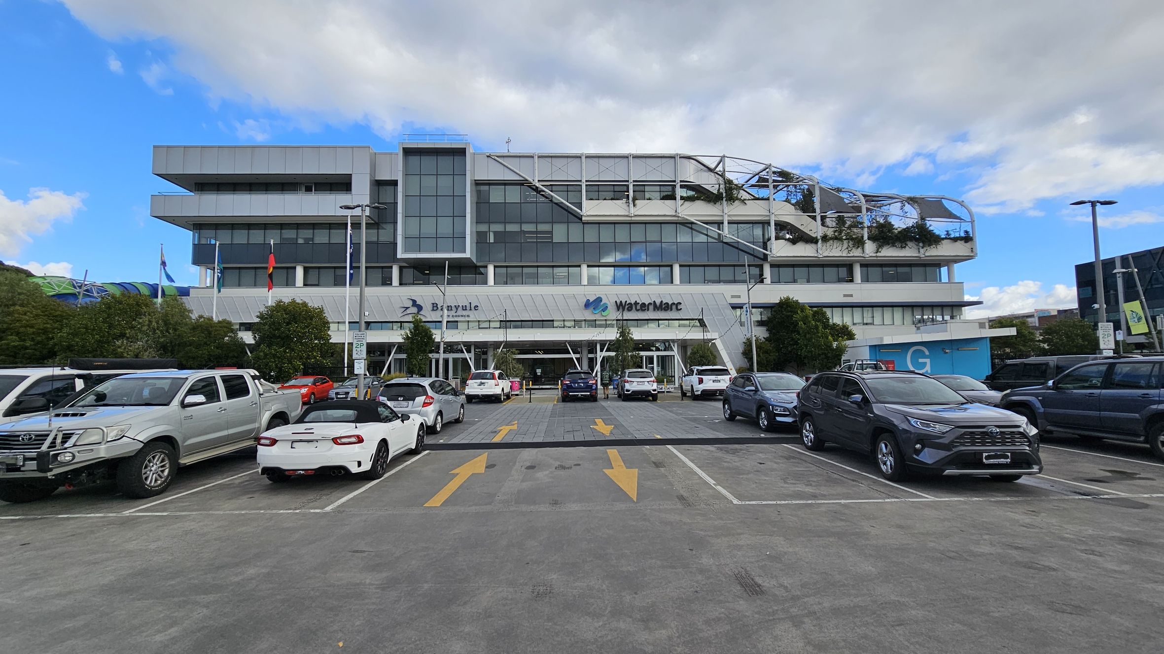 A multi-storey building with a modern design, featuring large glass windows and a rooftop garden with greenery. The building has signs for "Banyule" and "WaterMarc" on the front. In the foreground, a car park with various vehicles, including a white convertible and a silver pickup truck. Yellow arrows are painted on the asphalt, indicating traffic direction. Trees and flagpoles are positioned near the entrance. The sky is partly cloudy. A multi-storey building with a modern design, featuring large glass windows and a rooftop garden with greenery. The building has signs for "Banyule" and "WaterMarc" on the front. In the foreground, a car park with various vehicles, including a white convertible and a silver pickup truck. Yellow arrows are painted on the asphalt, indicating traffic direction. Trees and flagpoles are positioned near the entrance. The sky is partly cloudy.