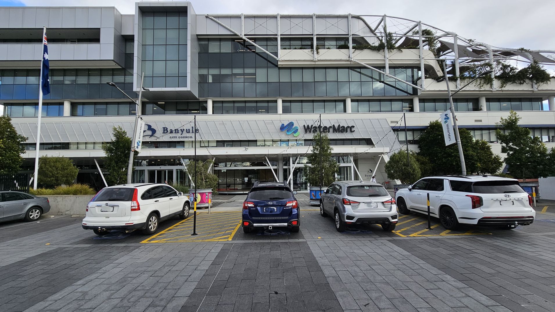 A modern multi-storey building with large glass windows and a geometric design. In the foreground, there are four parked cars on a paved area. The building features signage for "Banyule City Council" and "WaterMarc" above the entrance. There are several trees and a flagpole with a flag on the left. The structure has a distinctive roof with greenery and metal framework. The pavement is grey with a pattern of rectangular tiles. A modern multi-storey building with large glass windows and a geometric design. In the foreground, there are four parked cars on a paved area. The building features signage for "Banyule City Council" and "WaterMarc" above the entrance. There are several trees and a flagpole with a flag on the left. The structure has a distinctive roof with greenery and metal framework. The pavement is grey with a pattern of rectangular tiles.
