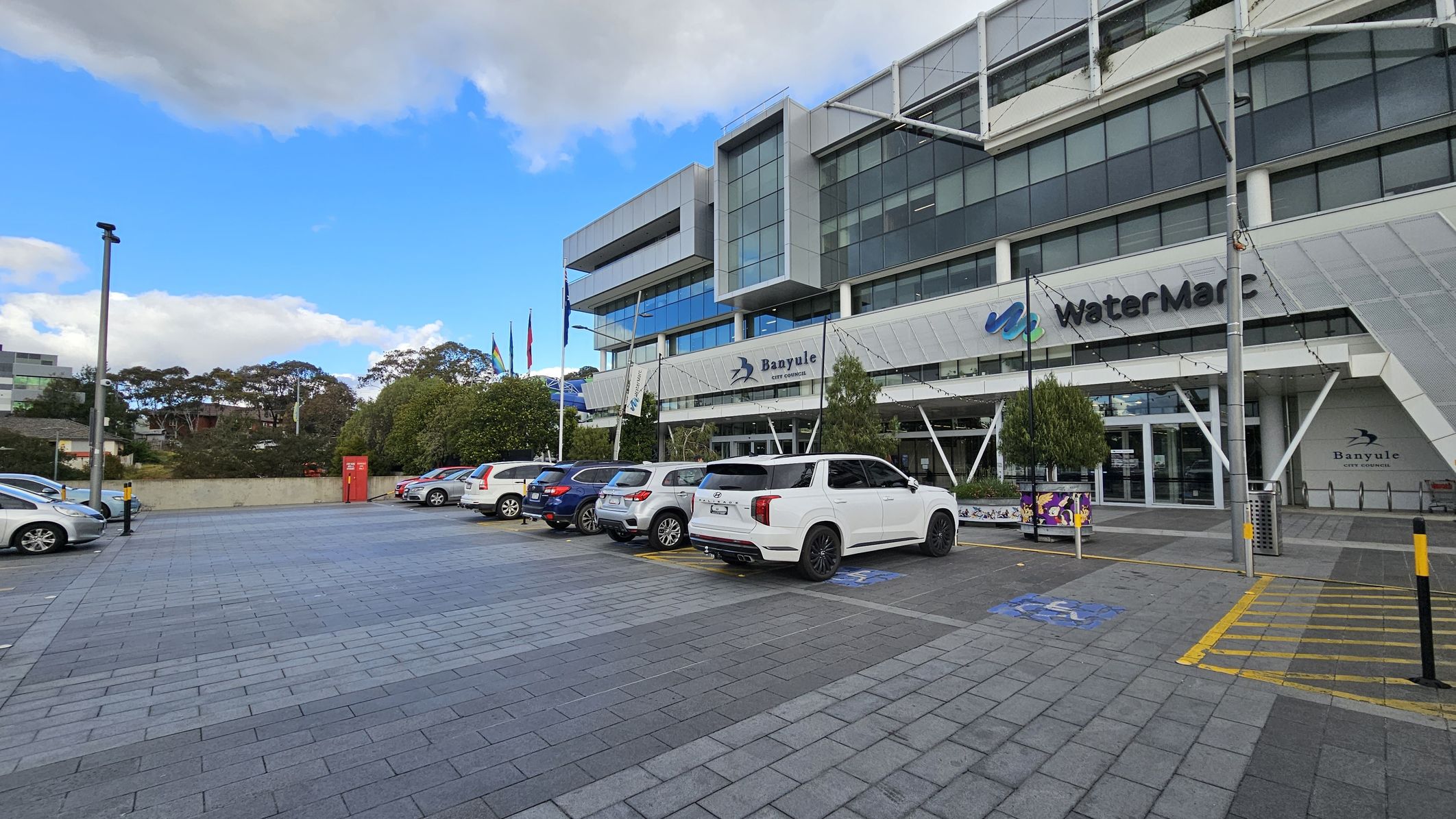A modern building with large glass windows and a sign reading "WaterMarc" and "Banyule City Council" on the facade. Several cars are parked in the foreground on a paved area. Flags are visible near the entrance, and there are trees and bushes along the side. A red utility box is positioned near the centre-left. The building has a sleek, angular design with metal and glass elements. A modern building with large glass windows and a sign reading "WaterMarc" and "Banyule City Council" on the facade. Several cars are parked in the foreground on a paved area. Flags are visible near the entrance, and there are trees and bushes along the side. A red utility box is positioned near the centre-left. The building has a sleek, angular design with metal and glass elements.