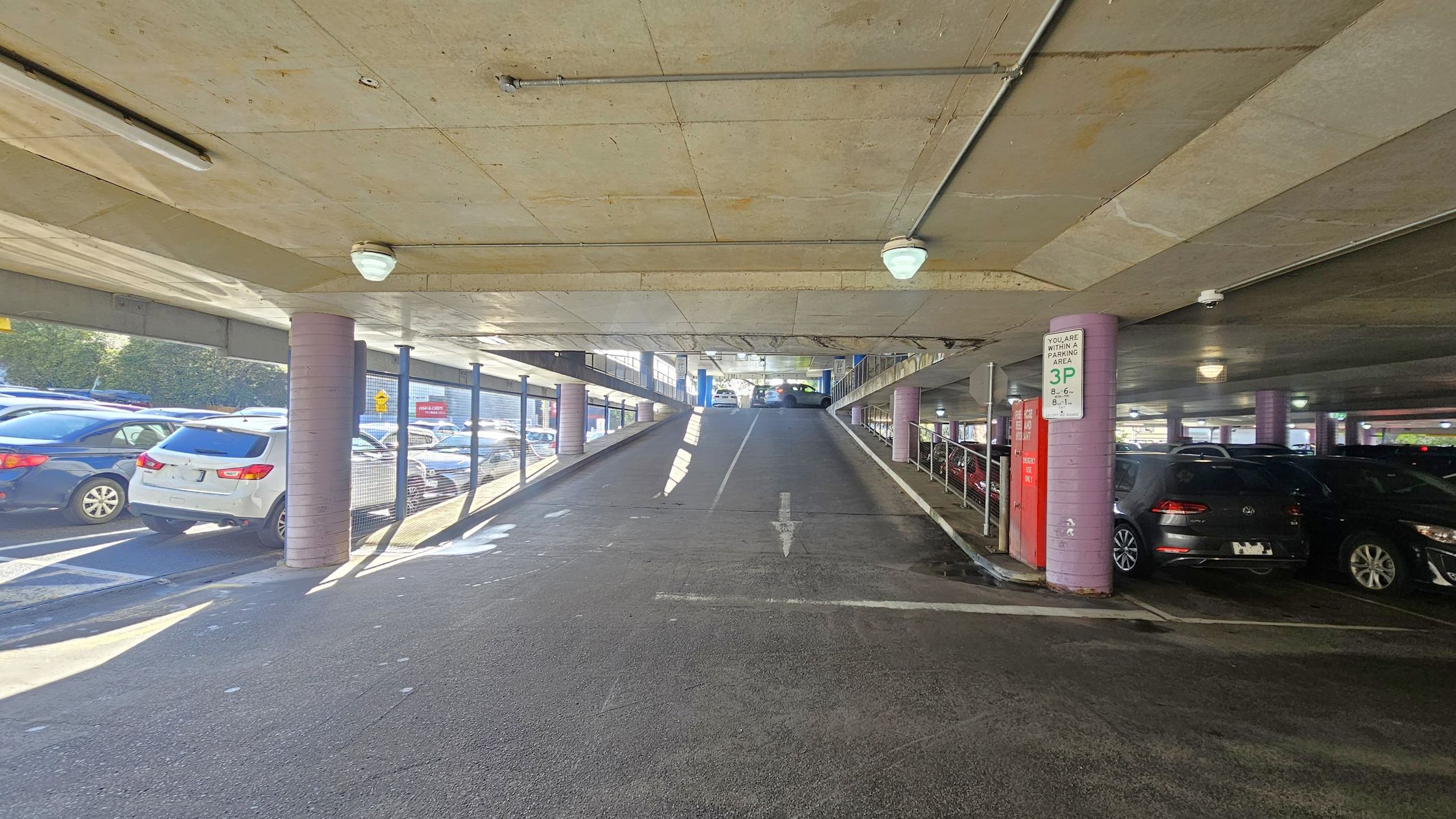 A multi-storey car park with a ramp in the centre leading upwards. On the left, several parked cars are visible, including a white SUV. On the right, more cars are parked, with a sign indicating parking restrictions. The ceiling is concrete with lights, and the floor is asphalt. Pink cylindrical columns support the structure, and metal railings line the ramp. Natural light filters in from the left side, illuminating the area. A multi-storey car park with a ramp in the centre leading upwards. On the left, several parked cars are visible, including a white SUV. On the right, more cars are parked, with a sign indicating parking restrictions. The ceiling is concrete with lights, and the floor is asphalt. Pink cylindrical columns support the structure, and metal railings line the ramp. Natural light filters in from the left side, illuminating the area.