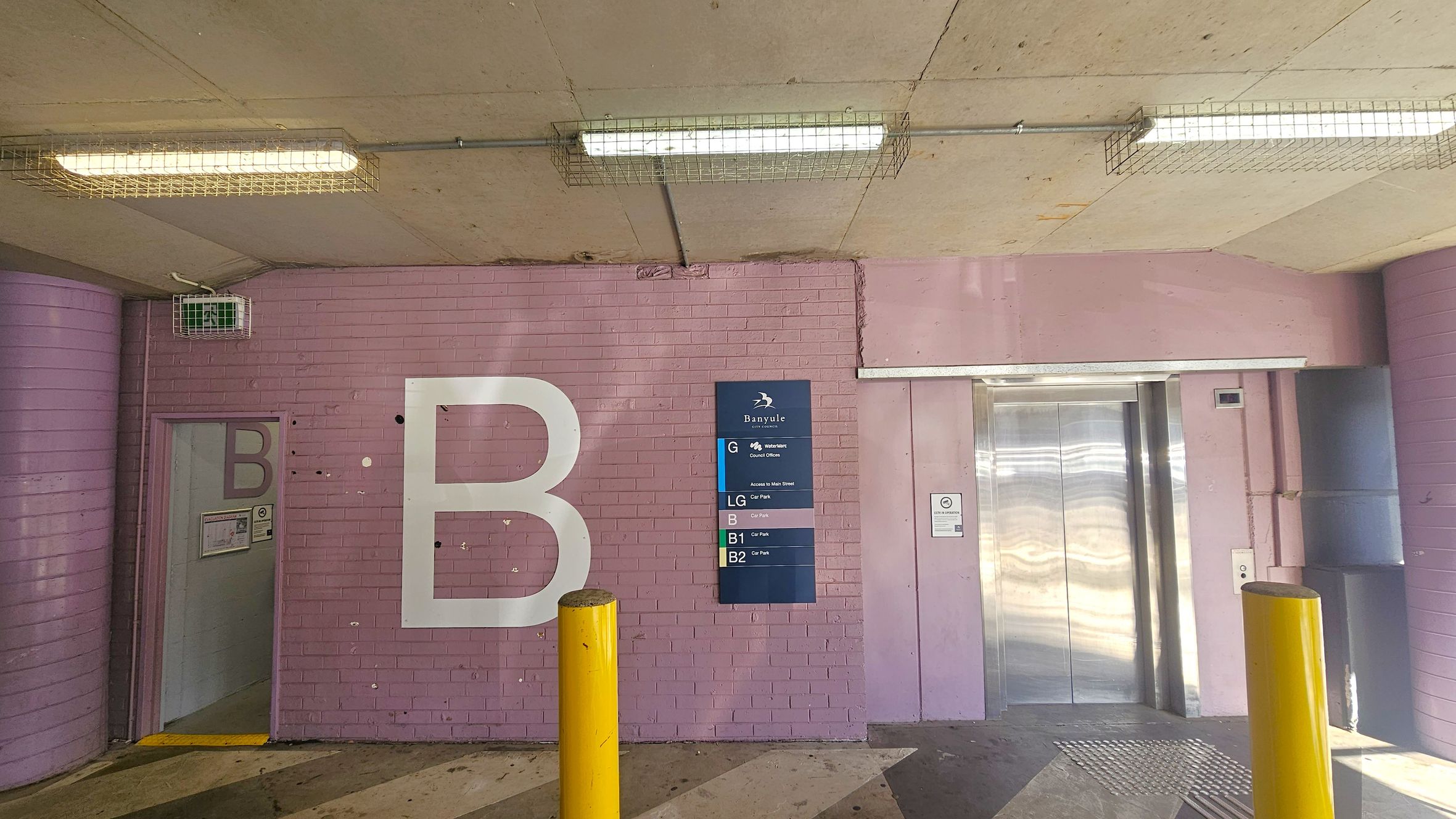 A pink brick wall with a large white letter "B" on the left. A small door with another "B" is partially open. In the centre, a blue sign lists different levels of a building. On the right, there is a metallic lift with closed doors. Two yellow bollards are in front of the wall. The ceiling is concrete with fluorescent lights encased in wire cages. The floor has a striped pattern. A pink brick wall with a large white letter "B" on the left. A small door with another "B" is partially open. In the centre, a blue sign lists different levels of a building. On the right, there is a metallic lift with closed doors. Two yellow bollards are in front of the wall. The ceiling is concrete with fluorescent lights encased in wire cages. The floor has a striped pattern.