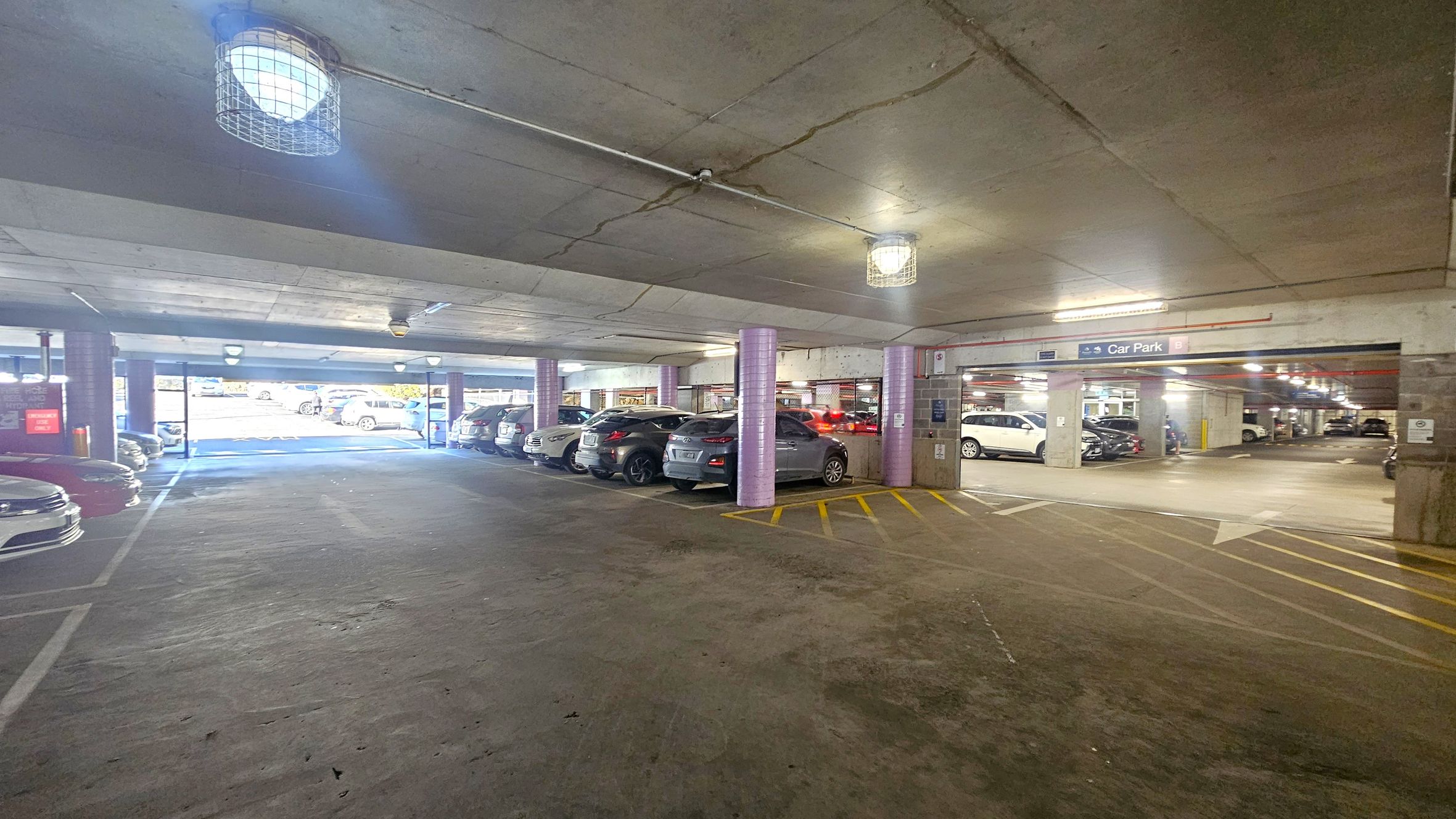 A multi-storey car park with concrete floors and ceilings. On the left, several parked cars are visible, with a bright light coming from the entrance. The right side shows more parked cars and a sign indicating "Car Park B". The ceiling has exposed lighting fixtures with protective cages. The floor has yellow markings and arrows guiding traffic flow. The area is well-lit with natural light from the entrance and artificial lights overhead. A multi-storey car park with concrete floors and ceilings. On the left, several parked cars are visible, with a bright light coming from the entrance. The right side shows more parked cars and a sign indicating "Car Park B". The ceiling has exposed lighting fixtures with protective cages. The floor has yellow markings and arrows guiding traffic flow. The area is well-lit with natural light from the entrance and artificial lights overhead.