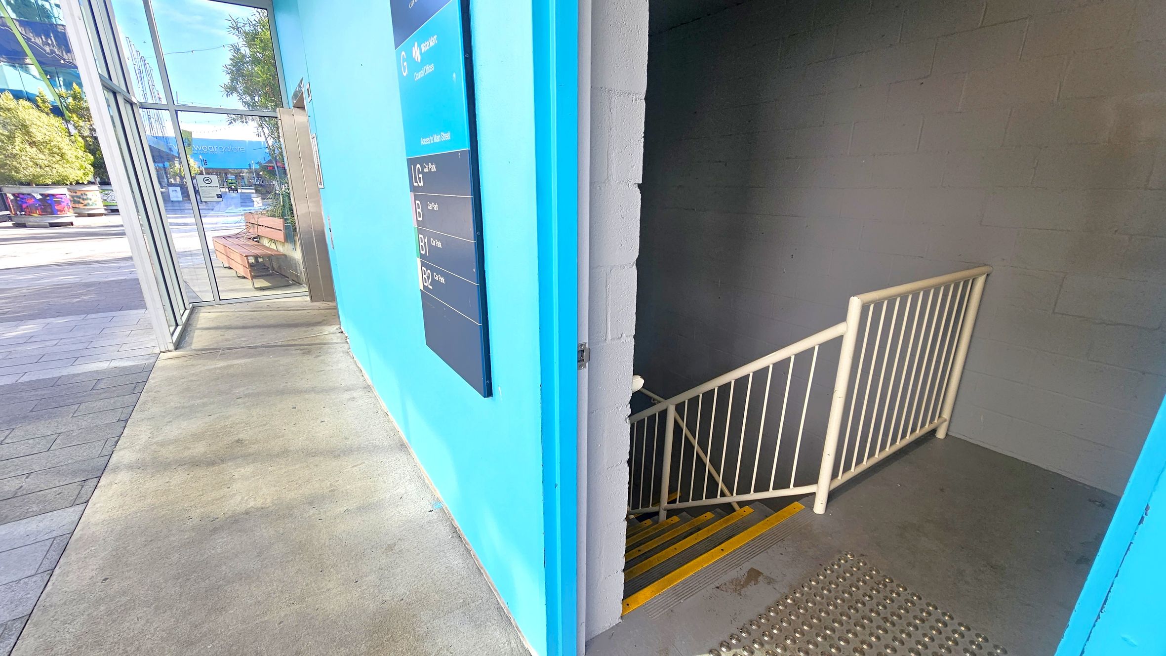 On the left, there's a glass entrance with a view of an outdoor area featuring benches and trees. The floor is a mix of concrete and tiles. On the right, a stairwell with a metal railing descends, marked by yellow safety strips. The wall is painted bright blue with a directory sign listing different levels. The ceiling is not visible. On the left, there's a glass entrance with a view of an outdoor area featuring benches and trees. The floor is a mix of concrete and tiles. On the right, a stairwell with a metal railing descends, marked by yellow safety strips. The wall is painted bright blue with a directory sign listing different levels. The ceiling is not visible.