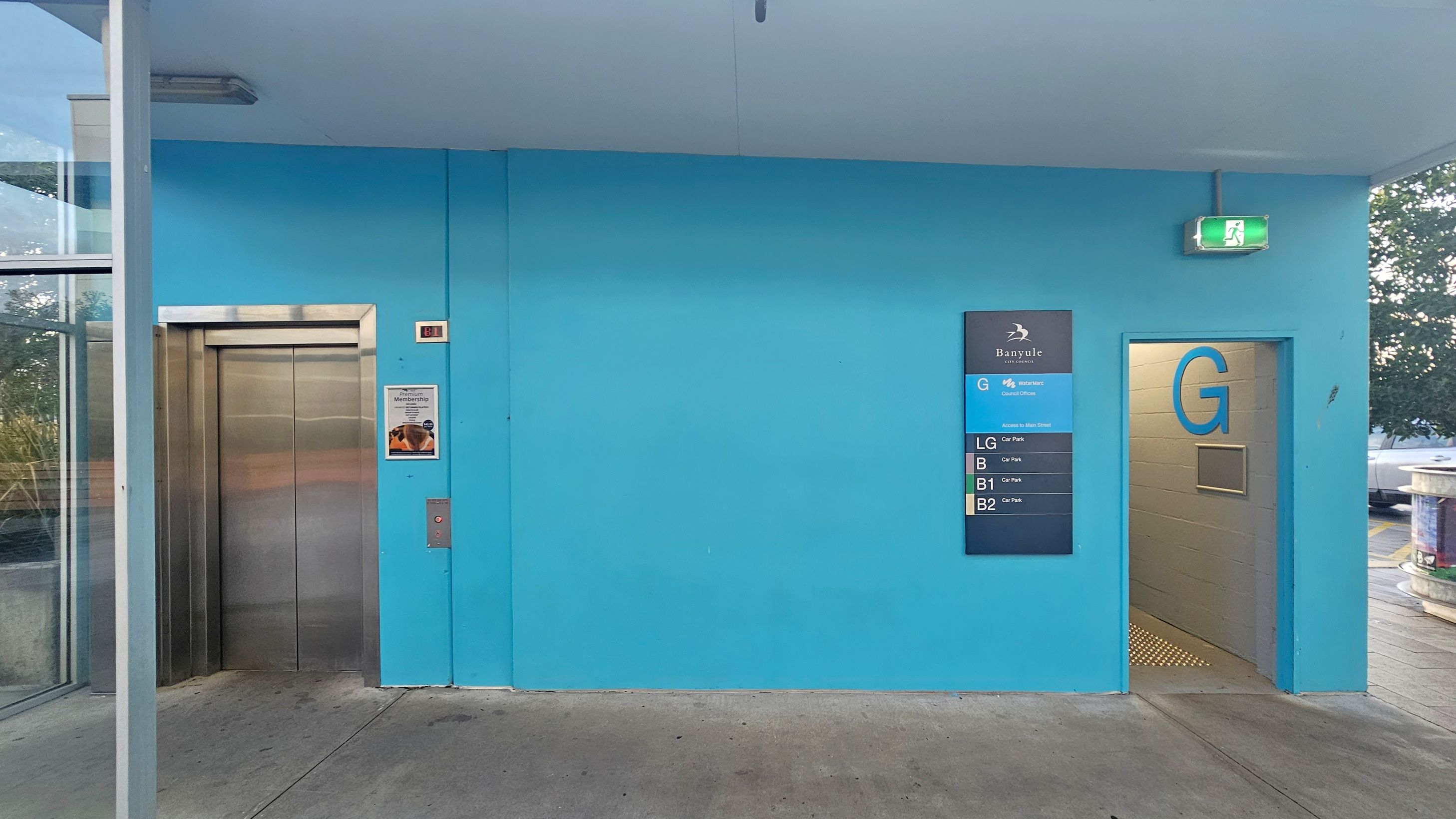 A blue wall with a lift on the left, featuring a stainless steel door and a poster beside it. To the right, a sign displays floor information for a building, labelled "Banyule" with levels G, LG, B, B1, and B2. An open doorway marked "G" leads to a corridor. Above the doorway, a green emergency exit sign is visible. The floor is concrete, and the ceiling is plain. A glass panel on the far left reveals some greenery outside. A blue wall with a lift on the left, featuring a stainless steel door and a poster beside it. To the right, a sign displays floor information for a building, labelled "Banyule" with levels G, LG, B, B1, and B2. An open doorway marked "G" leads to a corridor. Above the doorway, a green emergency exit sign is visible. The floor is concrete, and the ceiling is plain. A glass panel on the far left reveals some greenery outside.