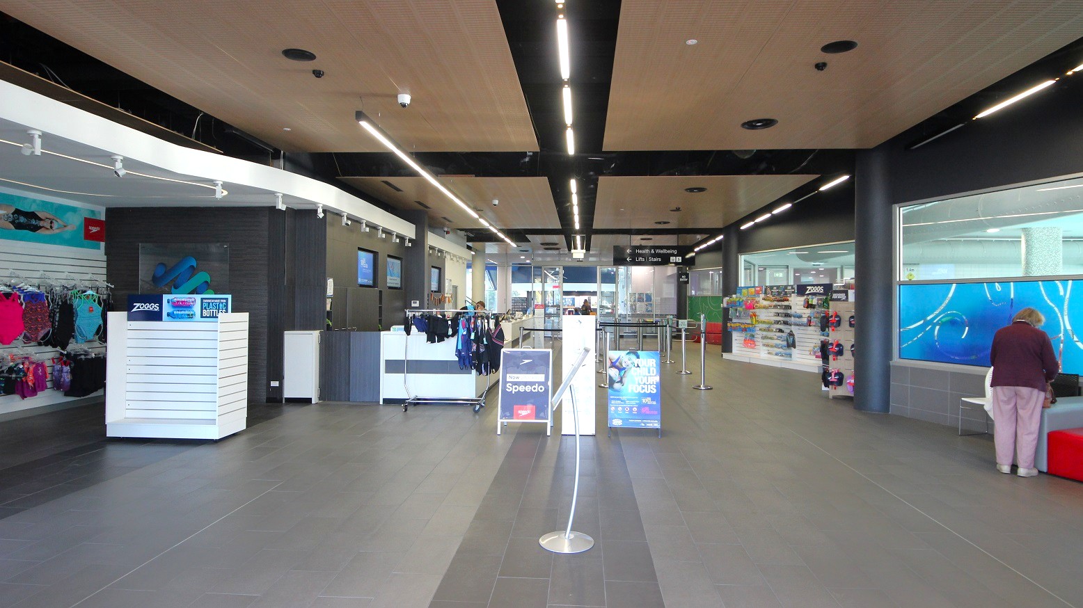 A spacious indoor area with a modern design. On the left, there are racks displaying swimwear and accessories. In the centre, a counter with hanging swim gear and promotional signs for Speedo. On the right, a person is standing near a bench, with a display of swimming goggles and accessories behind them. The floor is tiled in grey, and the ceiling features wooden panels with black beams and recessed lighting. A spacious indoor area with a modern design. On the left, there are racks displaying swimwear and accessories. In the centre, a counter with hanging swim gear and promotional signs for Speedo. On the right, a person is standing near a bench, with a display of swimming goggles and accessories behind them. The floor is tiled in grey, and the ceiling features wooden panels with black beams and recessed lighting.