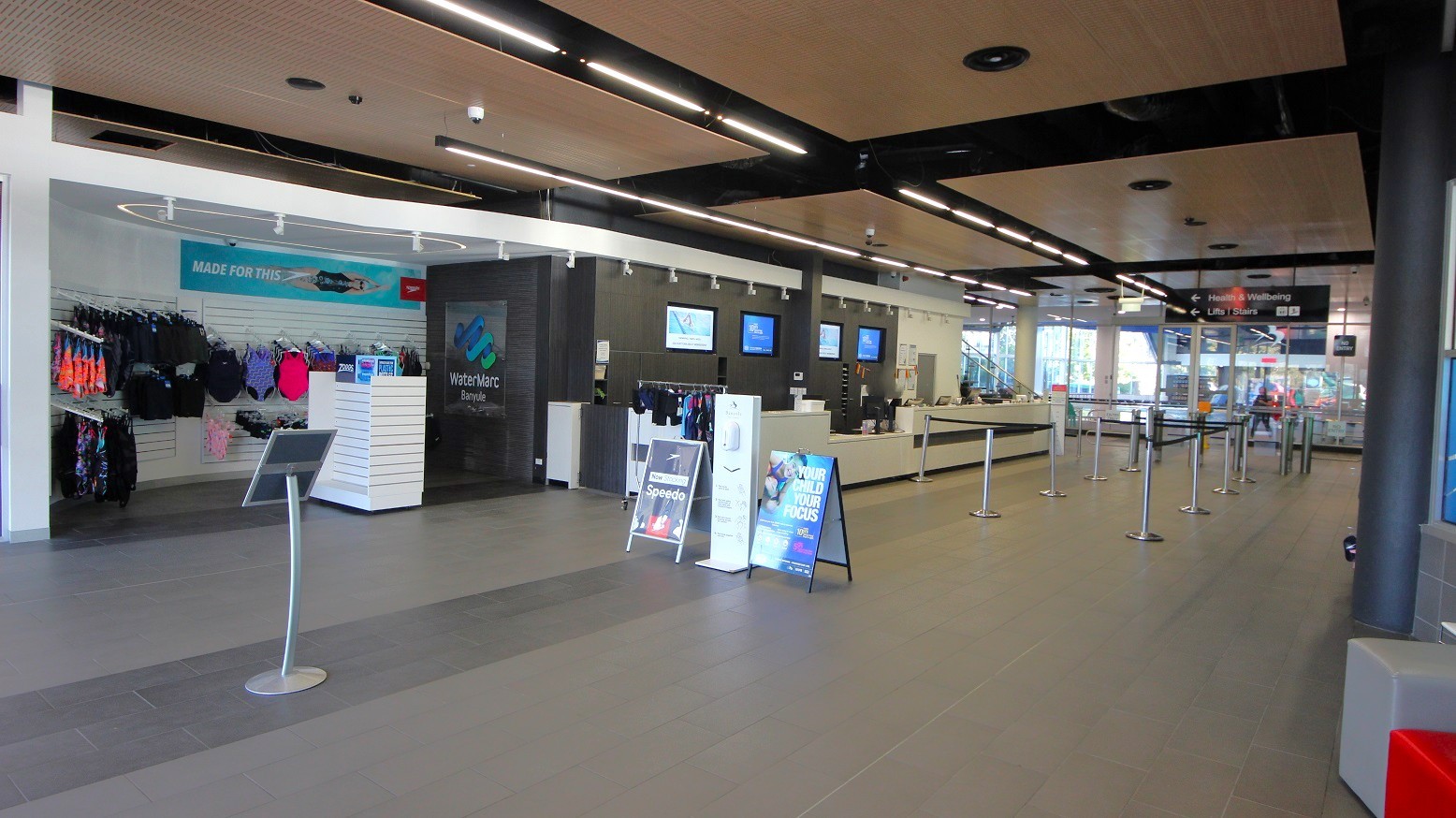 On the left, there are racks with swimwear and accessories against a white wall. In the centre, a stand with promotional signs is visible. To the right, a long reception counter with several monitors above it is present. The floor is a mix of grey and beige tiles, and the ceiling features wooden panels with black beams and recessed lighting. In the background, glass doors and windows allow natural light to enter the space. On the left, there are racks with swimwear and accessories against a white wall. In the centre, a stand with promotional signs is visible. To the right, a long reception counter with several monitors above it is present. The floor is a mix of grey and beige tiles, and the ceiling features wooden panels with black beams and recessed lighting. In the background, glass doors and windows allow natural light to enter the space.