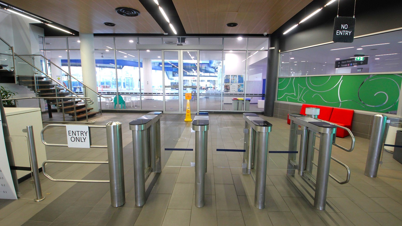 A modern indoor entrance area with turnstiles in the foreground, marked "ENTRY ONLY". To the left, a staircase with metal railings leads upwards. The background features glass doors and windows, revealing a brightly lit interior space. On the right, a green wall with abstract patterns and a red bench. The ceiling has recessed lighting and ventilation fixtures. A "NO ENTRY" sign hangs above the turnstiles. The floor is tiled in a neutral colour. A modern indoor entrance area with turnstiles in the foreground, marked "ENTRY ONLY". To the left, a staircase with metal railings leads upwards. The background features glass doors and windows, revealing a brightly lit interior space. On the right, a green wall with abstract patterns and a red bench. The ceiling has recessed lighting and ventilation fixtures. A "NO ENTRY" sign hangs above the turnstiles. The floor is tiled in a neutral colour.