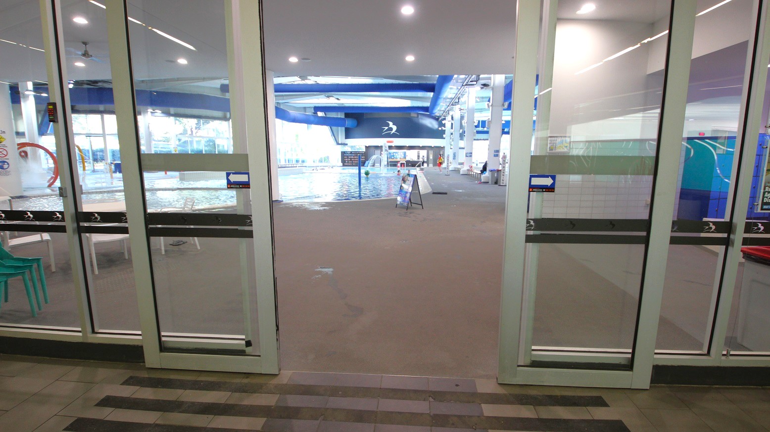Glass doors open to an indoor swimming pool area. On the left, there are stacked green chairs and a glimpse of a play area with a red circular feature. In the centre, the pool is visible with a signboard on the poolside. The right side shows a blue and white wall with a design. The ceiling has recessed lighting and blue structural beams. The floor is tiled in a light brown colour. Glass doors open to an indoor swimming pool area. On the left, there are stacked green chairs and a glimpse of a play area with a red circular feature. In the centre, the pool is visible with a signboard on the poolside. The right side shows a blue and white wall with a design. The ceiling has recessed lighting and blue structural beams. The floor is tiled in a light brown colour.