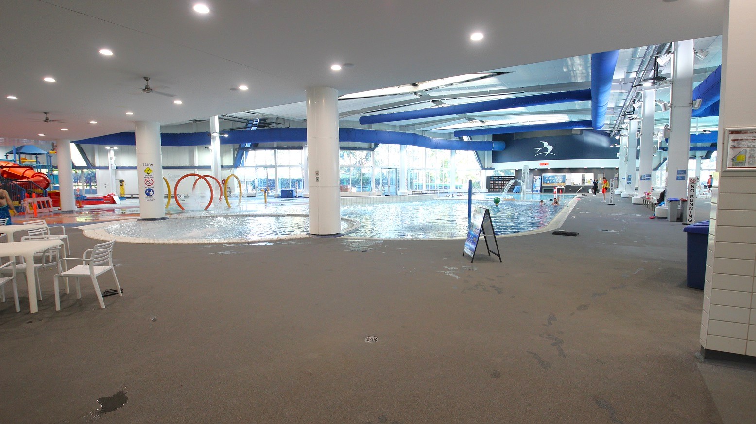Indoor swimming pool area with a large pool in the centre. On the left, there are white tables and chairs, and a red water slide. In the background, large windows allow natural light to enter. The ceiling is white with blue ducts and recessed lighting. A signboard is placed near the pool, and there are various water play features like hoops. The floor is wet and made of non-slip material. Indoor swimming pool area with a large pool in the centre. On the left, there are white tables and chairs, and a red water slide. In the background, large windows allow natural light to enter. The ceiling is white with blue ducts and recessed lighting. A signboard is placed near the pool, and there are various water play features like hoops. The floor is wet and made of non-slip material.