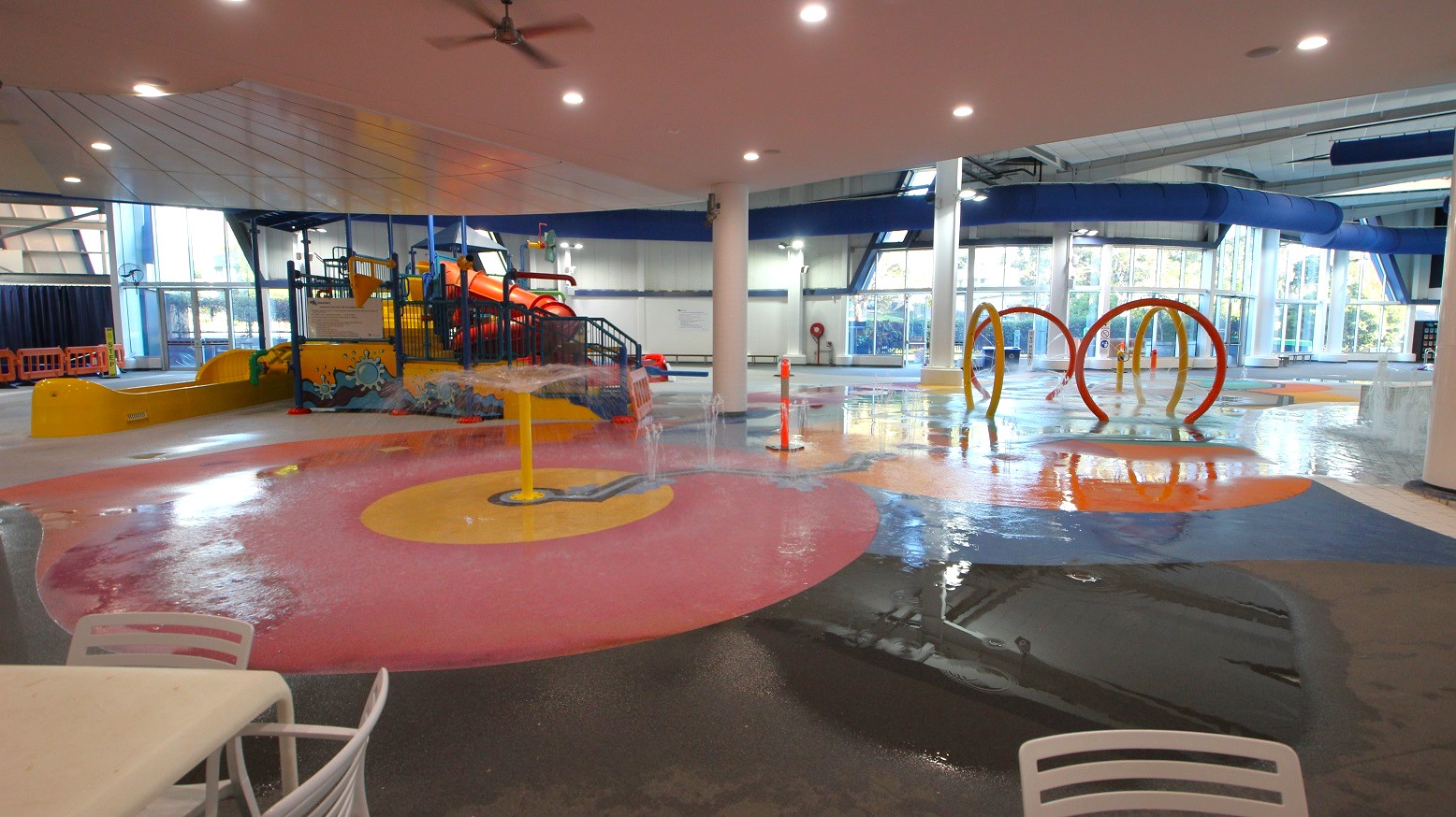 Indoor water play area with colourful features. On the left, a large play structure with slides in orange and yellow. In the centre, a pink and yellow splash pad with water jets. On the right, orange and yellow hoops for water play. The floor is wet and reflective, with a white table and chairs in the foreground. The ceiling is white with recessed lighting, and large windows in the background allow natural light in. Indoor water play area with colourful features. On the left, a large play structure with slides in orange and yellow. In the centre, a pink and yellow splash pad with water jets. On the right, orange and yellow hoops for water play. The floor is wet and reflective, with a white table and chairs in the foreground. The ceiling is white with recessed lighting, and large windows in the background allow natural light in.