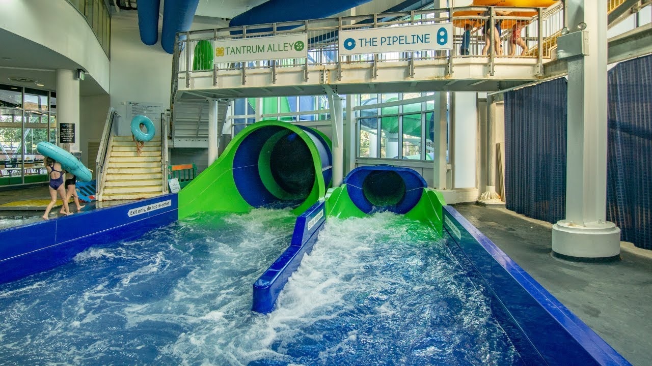 Indoor water park with two large water slides in the centre, one green and one blue. The slides empty into a pool with churning water. On the left, two people carry inflatable tubes up a staircase. Above the slides, signs read "Tantrum Alley" and "The Pipeline." The area is enclosed with large windows in the background, allowing natural light. The floor is wet and grey, and the ceiling has exposed blue pipes. Indoor water park with two large water slides in the centre, one green and one blue. The slides empty into a pool with churning water. On the left, two people carry inflatable tubes up a staircase. Above the slides, signs read "Tantrum Alley" and "The Pipeline." The area is enclosed with large windows in the background, allowing natural light. The floor is wet and grey, and the ceiling has exposed blue pipes.