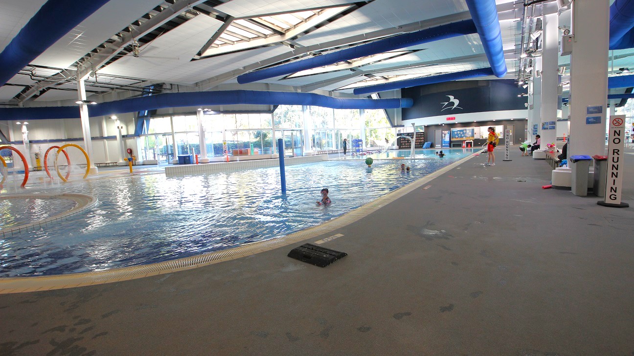 An indoor swimming pool area with a large pool in the centre. On the left, there are colourful hoops emerging from the water. The right side features a wide walkway with a "No Running" sign and several people sitting on benches. The ceiling is high with exposed beams and blue ducts. Large windows in the background allow natural light to enter, and a lifeguard stands near the pool. The floor is grey and textured. An indoor swimming pool area with a large pool in the centre. On the left, there are colourful hoops emerging from the water. The right side features a wide walkway with a "No Running" sign and several people sitting on benches. The ceiling is high with exposed beams and blue ducts. Large windows in the background allow natural light to enter, and a lifeguard stands near the pool. The floor is grey and textured.