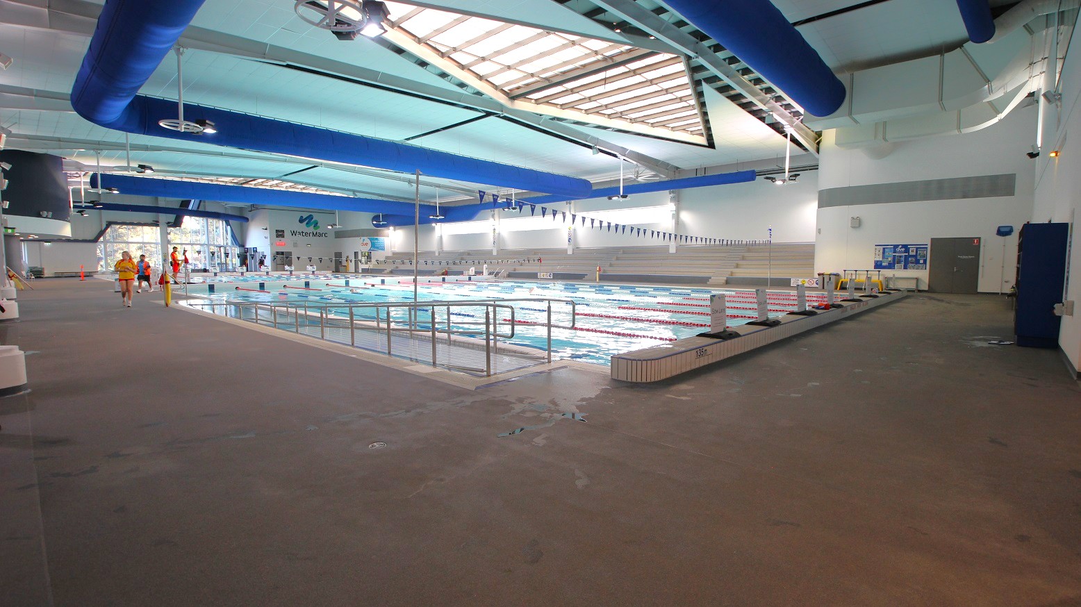 Indoor swimming pool with multiple lanes, surrounded by a wide deck area. On the left, a few people in swimwear and lifeguard attire walk along the deck. The pool is in the centre, with lane dividers and a railing. The ceiling features large blue ventilation ducts and skylights, allowing natural light. The walls are white with some signage, and there is a door on the right. The floor is a non-slip surface, and the area is well-lit. Indoor swimming pool with multiple lanes, surrounded by a wide deck area. On the left, a few people in swimwear and lifeguard attire walk along the deck. The pool is in the centre, with lane dividers and a railing. The ceiling features large blue ventilation ducts and skylights, allowing natural light. The walls are white with some signage, and there is a door on the right. The floor is a non-slip surface, and the area is well-lit.