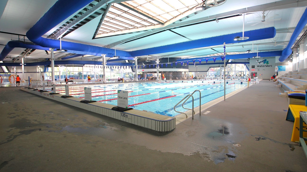 An indoor swimming pool with multiple lanes, separated by red and white lane dividers. On the left, there are lifeguards in orange vests near the pool's edge. The right side features a tiled pool deck with a metal handrail leading into the water. Blue ducts run along the ceiling, which has a large skylight. The floor is wet and textured for safety. Benches and a sign reading "WaterMarc" are visible in the background. An indoor swimming pool with multiple lanes, separated by red and white lane dividers. On the left, there are lifeguards in orange vests near the pool's edge. The right side features a tiled pool deck with a metal handrail leading into the water. Blue ducts run along the ceiling, which has a large skylight. The floor is wet and textured for safety. Benches and a sign reading "WaterMarc" are visible in the background.