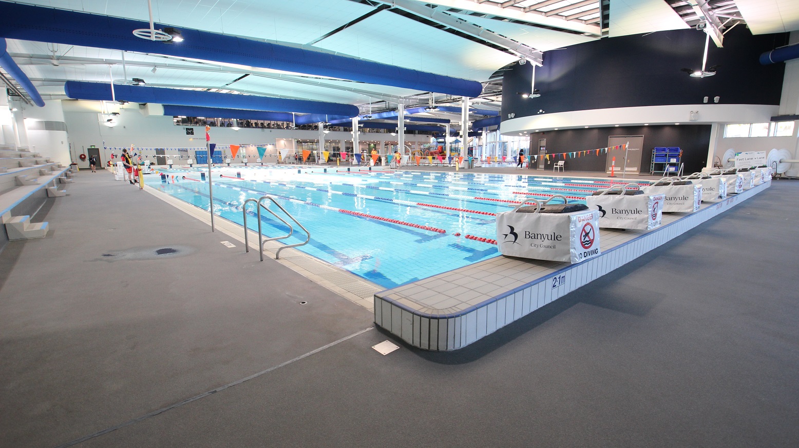 An indoor swimming pool with multiple lanes, separated by red lane dividers. On the left, there are tiered seating areas with grey benches. The pool deck is grey, with a metal handrail leading into the water. On the right, there are signs with "Banyule" and "No Diving" warnings. The ceiling features blue and white panels with exposed beams and lighting fixtures. In the background, there are people near the pool and a dark wall with windows above. An indoor swimming pool with multiple lanes, separated by red lane dividers. On the left, there are tiered seating areas with grey benches. The pool deck is grey, with a metal handrail leading into the water. On the right, there are signs with "Banyule" and "No Diving" warnings. The ceiling features blue and white panels with exposed beams and lighting fixtures. In the background, there are people near the pool and a dark wall with windows above.