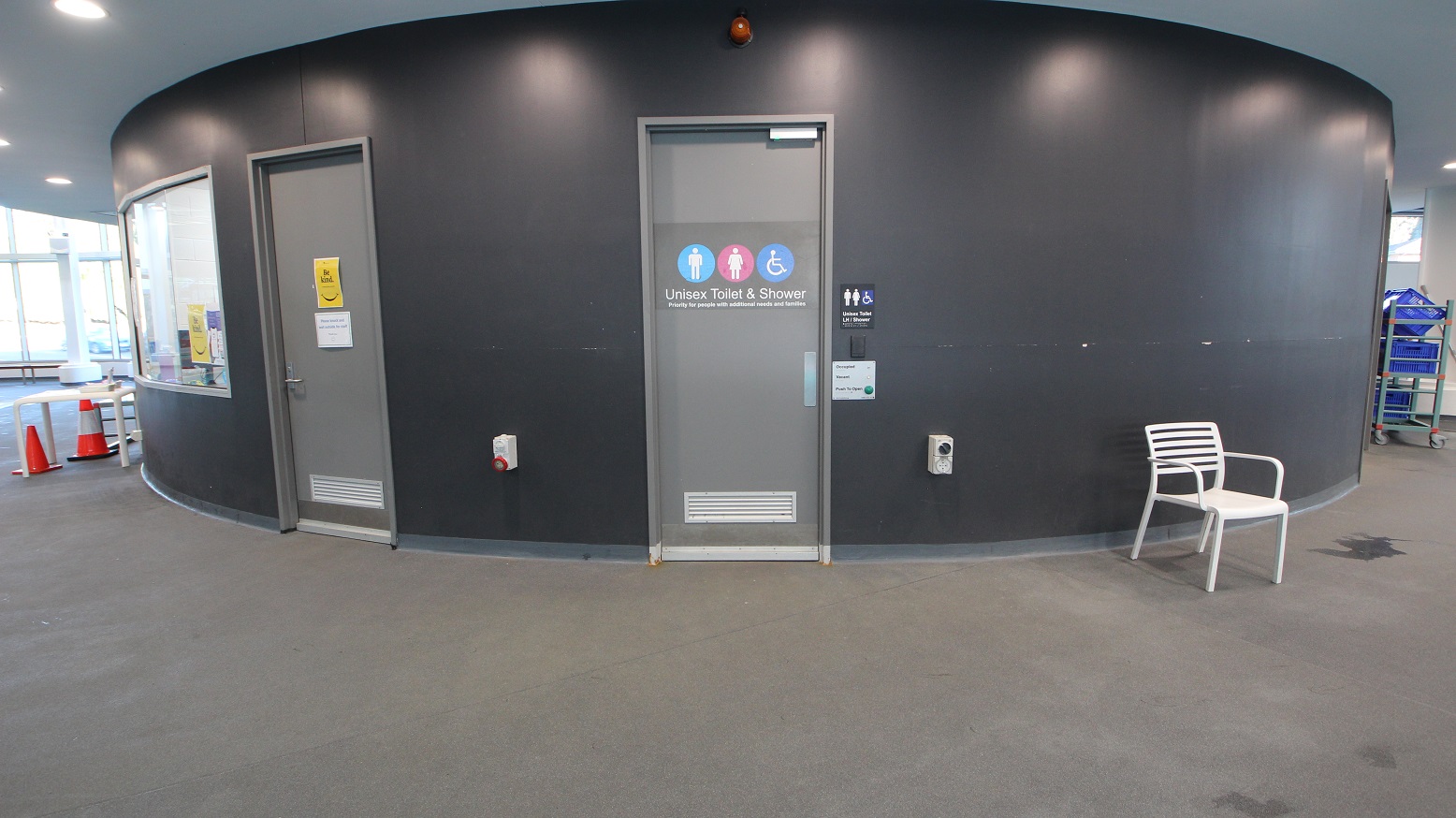 A curved, dark grey wall with two doors. The central door is marked "Unisex Toilet & Shower" with symbols for accessibility. To the left, another door has a yellow sign. A white chair is positioned on the right side of the image. The floor is carpeted in grey, and the ceiling is light-coloured with recessed lighting. On the far left, there are orange traffic cones and a window, while a blue trolley is partially visible on the right. A curved, dark grey wall with two doors. The central door is marked "Unisex Toilet & Shower" with symbols for accessibility. To the left, another door has a yellow sign. A white chair is positioned on the right side of the image. The floor is carpeted in grey, and the ceiling is light-coloured with recessed lighting. On the far left, there are orange traffic cones and a window, while a blue trolley is partially visible on the right.