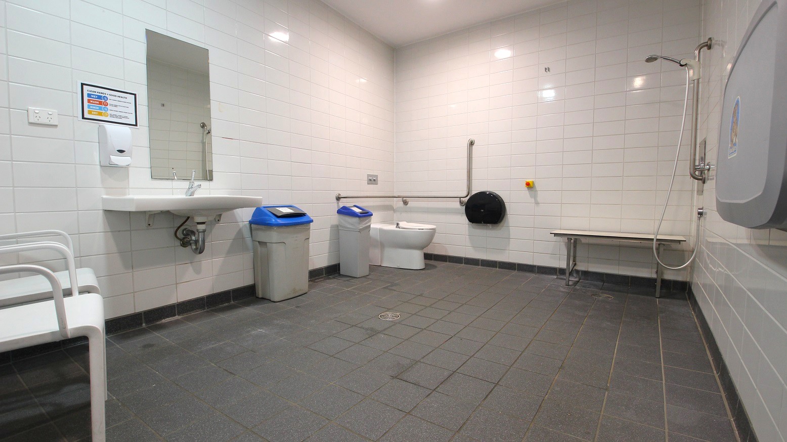 A spacious, accessible bathroom with white tiled walls and a dark tiled floor. On the left, there's a white sink with a mirror above it and a soap dispenser. Next to the sink are two bins with blue lids. In the centre, a toilet with a grab rail is visible. On the right, there's a shower area with a handheld showerhead and a foldable bench. A baby changing station is mounted on the wall. The ceiling is plain with recessed lighting. A spacious, accessible bathroom with white tiled walls and a dark tiled floor. On the left, there's a white sink with a mirror above it and a soap dispenser. Next to the sink are two bins with blue lids. In the centre, a toilet with a grab rail is visible. On the right, there's a shower area with a handheld showerhead and a foldable bench. A baby changing station is mounted on the wall. The ceiling is plain with recessed lighting.