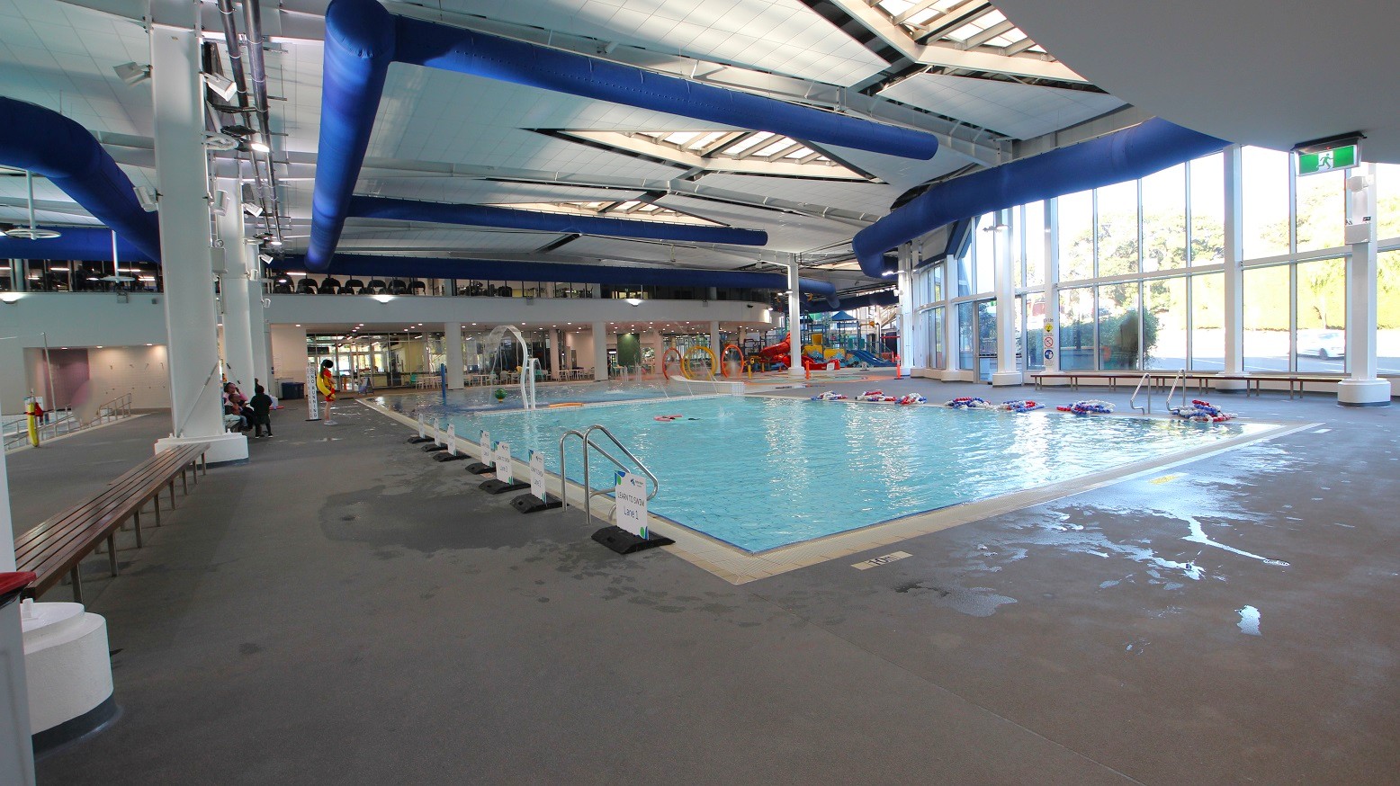 An indoor swimming pool area with a large pool on the right, surrounded by a tiled deck. Several flotation devices are floating in the water. On the left, there are benches and a few people seated. The ceiling features blue ventilation ducts and skylights, allowing natural light to illuminate the space. Large windows on the right wall provide additional light and a view outside. The floor is wet in places, indicating recent use. An indoor swimming pool area with a large pool on the right, surrounded by a tiled deck. Several flotation devices are floating in the water. On the left, there are benches and a few people seated. The ceiling features blue ventilation ducts and skylights, allowing natural light to illuminate the space. Large windows on the right wall provide additional light and a view outside. The floor is wet in places, indicating recent use.