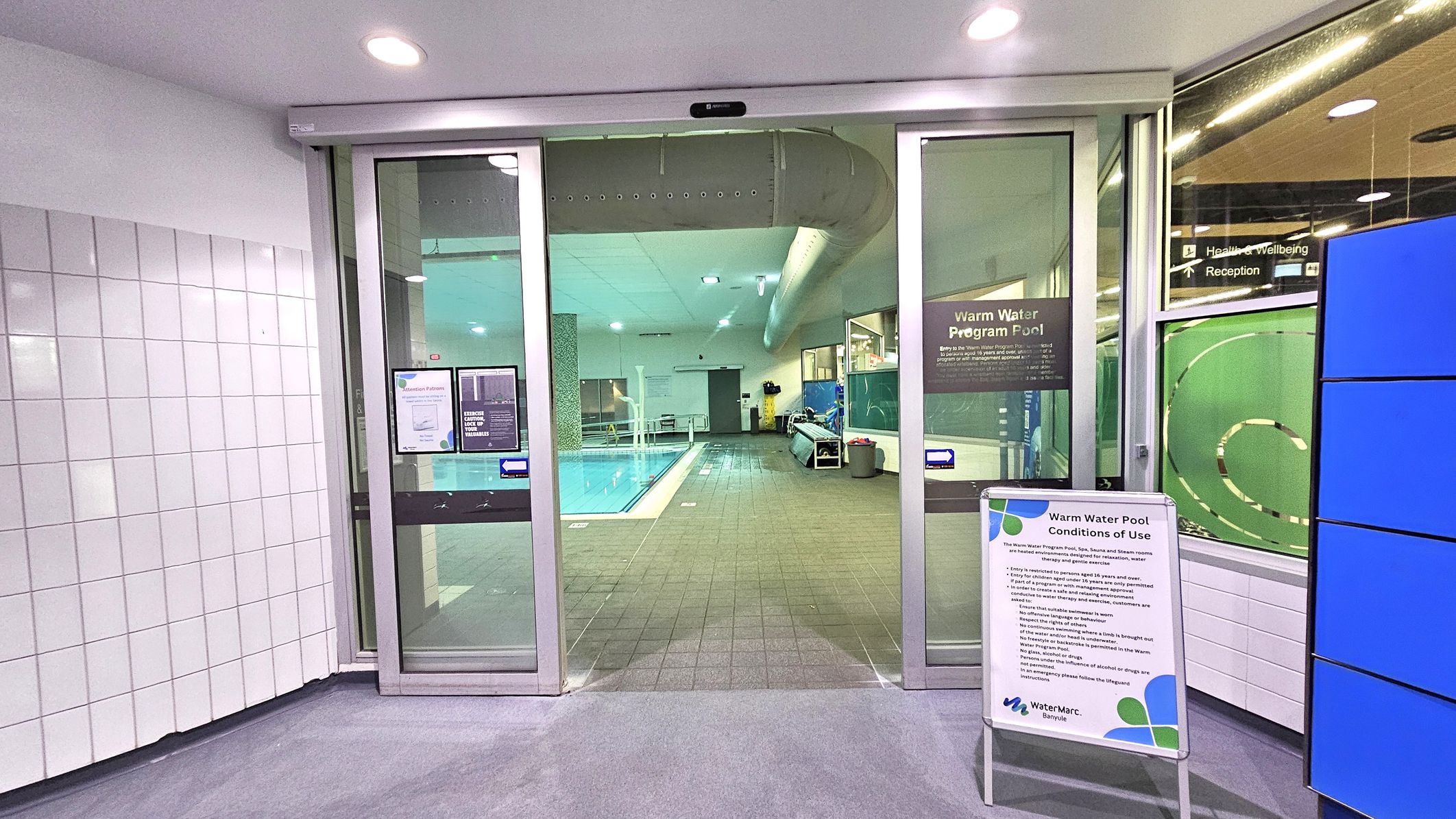 Sliding glass doors lead to an indoor pool area. The pool is visible in the centre, with a tiled floor surrounding it. On the left, white tiled walls are present. On the right, a sign with "Warm Water Pool Conditions of Use" is displayed. The ceiling has recessed lighting, and a large ventilation duct runs along it. In the background, pool equipment and a bench are visible. Sliding glass doors lead to an indoor pool area. The pool is visible in the centre, with a tiled floor surrounding it. On the left, white tiled walls are present. On the right, a sign with "Warm Water Pool Conditions of Use" is displayed. The ceiling has recessed lighting, and a large ventilation duct runs along it. In the background, pool equipment and a bench are visible.
