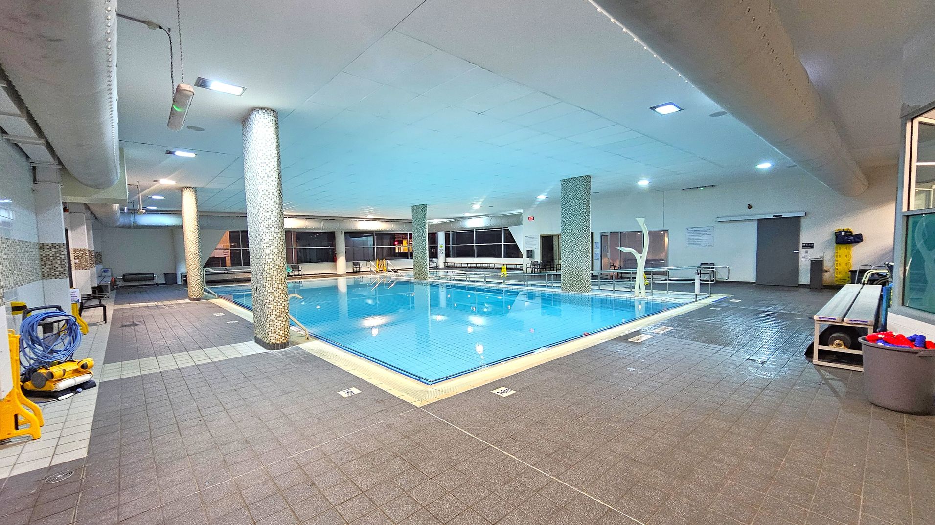 An indoor swimming pool with clear blue water is surrounded by a tiled floor. On the left, there are cleaning equipment and a coiled hose. The ceiling features recessed lighting and large ventilation ducts. In the background, large windows line the walls, and there are several benches and a door. A bin with red and blue items is on the right, next to a bench. The pool is bordered by mosaic-tiled columns. An indoor swimming pool with clear blue water is surrounded by a tiled floor. On the left, there are cleaning equipment and a coiled hose. The ceiling features recessed lighting and large ventilation ducts. In the background, large windows line the walls, and there are several benches and a door. A bin with red and blue items is on the right, next to a bench. The pool is bordered by mosaic-tiled columns.