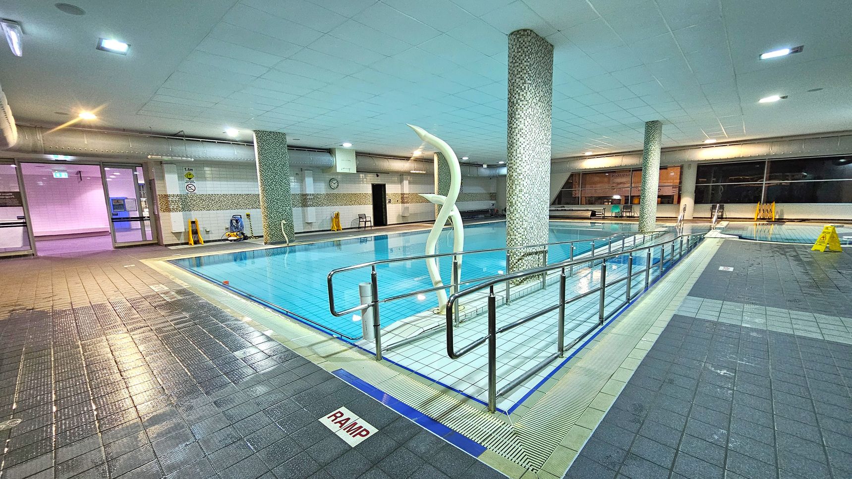 An indoor swimming pool with a tiled floor and ceiling. In the centre, a pool with clear blue water is surrounded by a metal railing. A decorative sculpture stands near a mosaic-tiled column. On the left, glass doors lead to another area, and a clock is visible on the wall. On the right, large windows and a few caution signs are present. The floor is wet, with a marked ramp area in the foreground. The lighting is bright and evenly distributed. An indoor swimming pool with a tiled floor and ceiling. In the centre, a pool with clear blue water is surrounded by a metal railing. A decorative sculpture stands near a mosaic-tiled column. On the left, glass doors lead to another area, and a clock is visible on the wall. On the right, large windows and a few caution signs are present. The floor is wet, with a marked ramp area in the foreground. The lighting is bright and evenly distributed.