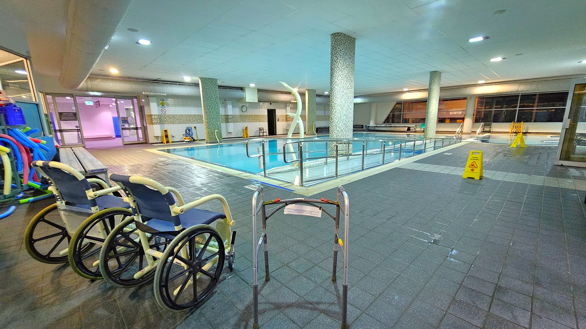 An indoor swimming pool area with a tiled floor and ceiling. On the left, there are two wheelchairs and a collection of pool noodles. In the centre, a pool is surrounded by a metal railing, with a decorative sculpture nearby. On the right, there are caution signs on the floor. Large windows line the background, and there are benches along the walls. The area is well-lit with overhead lights. An indoor swimming pool area with a tiled floor and ceiling. On the left, there are two wheelchairs and a collection of pool noodles. In the centre, a pool is surrounded by a metal railing, with a decorative sculpture nearby. On the right, there are caution signs on the floor. Large windows line the background, and there are benches along the walls. The area is well-lit with overhead lights.
