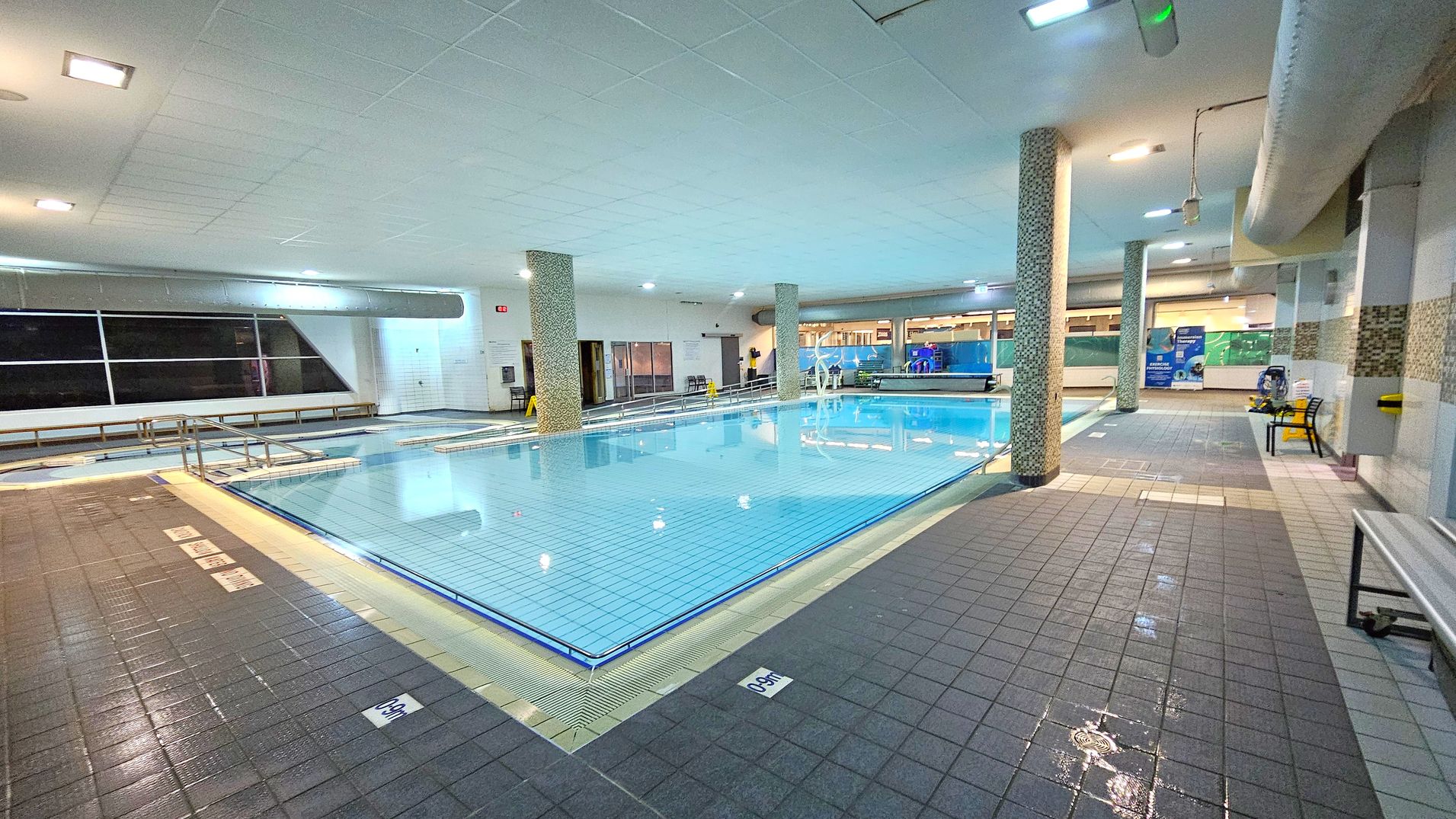 An indoor swimming pool with clear blue water is surrounded by a tiled deck. On the left, there are large windows and a row of benches. In the centre, two mosaic-tiled columns support the ceiling. On the right, there are informational posters and a yellow chair. The ceiling is white with recessed lighting, and the floor is covered with dark grey tiles, some of which are wet. A metal handrail leads into the pool. An indoor swimming pool with clear blue water is surrounded by a tiled deck. On the left, there are large windows and a row of benches. In the centre, two mosaic-tiled columns support the ceiling. On the right, there are informational posters and a yellow chair. The ceiling is white with recessed lighting, and the floor is covered with dark grey tiles, some of which are wet. A metal handrail leads into the pool.