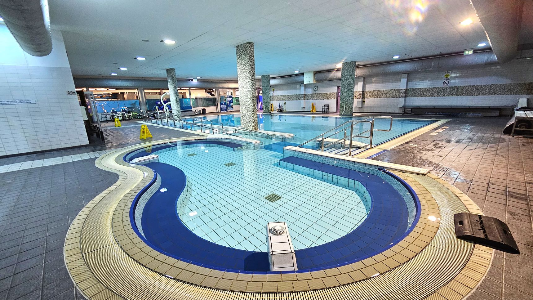 An indoor swimming pool area with a curvy, shallow pool in the foreground, surrounded by a beige and blue tiled border. The main pool is in the background, featuring handrails and a tiled deck. Several columns support the ceiling, which is fitted with recessed lighting. The floor is tiled in shades of grey and beige, with caution signs visible near the pool. Benches and a wall with a clock are on the right side. An indoor swimming pool area with a curvy, shallow pool in the foreground, surrounded by a beige and blue tiled border. The main pool is in the background, featuring handrails and a tiled deck. Several columns support the ceiling, which is fitted with recessed lighting. The floor is tiled in shades of grey and beige, with caution signs visible near the pool. Benches and a wall with a clock are on the right side.