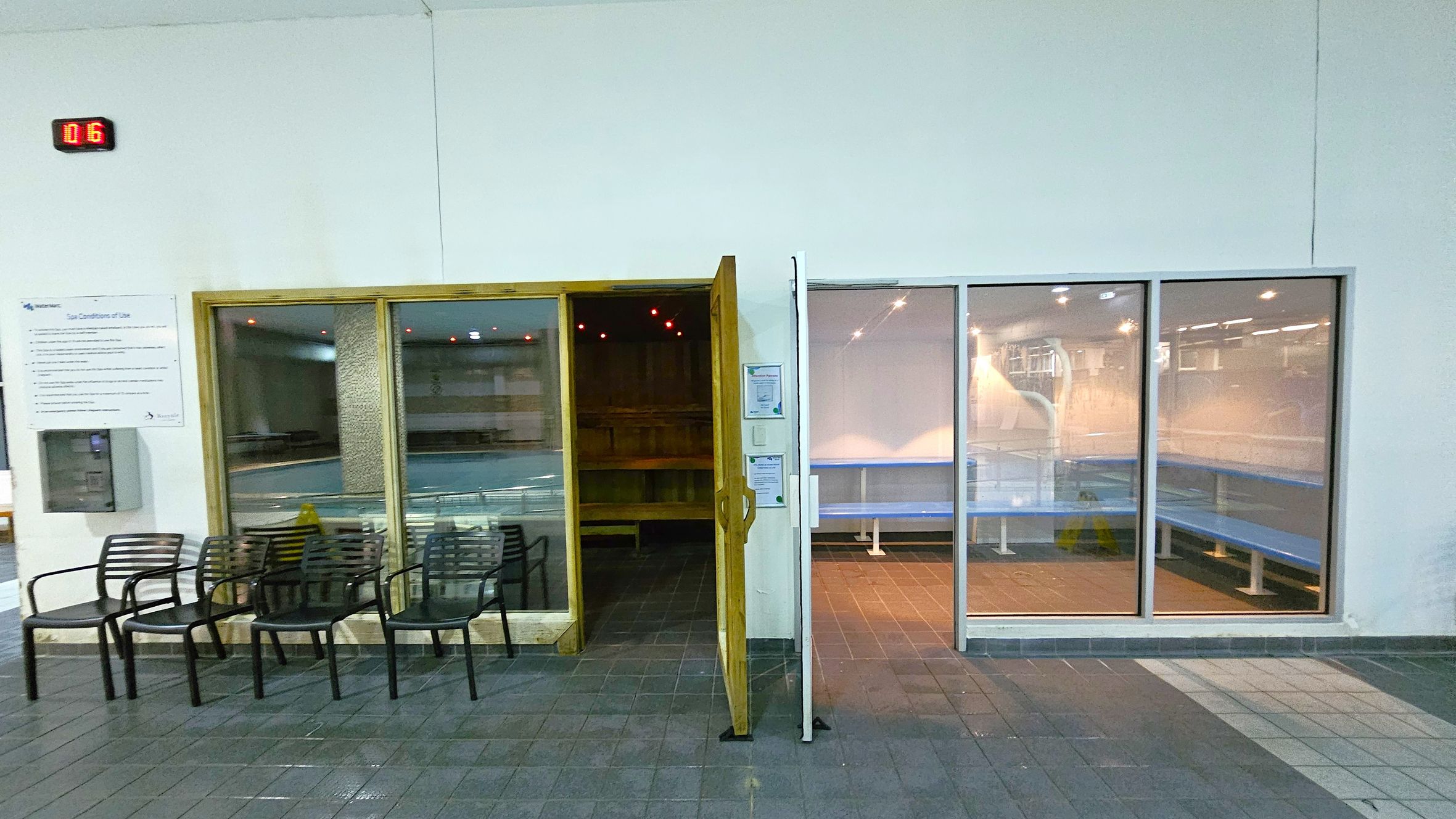 On the left, there are several black chairs lined up against a white wall. Above them, a digital display shows "016". A sign with text is mounted on the wall. In the centre, a wooden-framed glass door is open, revealing a dimly lit sauna with wooden benches. On the right, large glass panels show a room with blue benches and bright lighting. The floor is tiled in shades of grey and green. On the left, there are several black chairs lined up against a white wall. Above them, a digital display shows "016". A sign with text is mounted on the wall. In the centre, a wooden-framed glass door is open, revealing a dimly lit sauna with wooden benches. On the right, large glass panels show a room with blue benches and bright lighting. The floor is tiled in shades of grey and green.