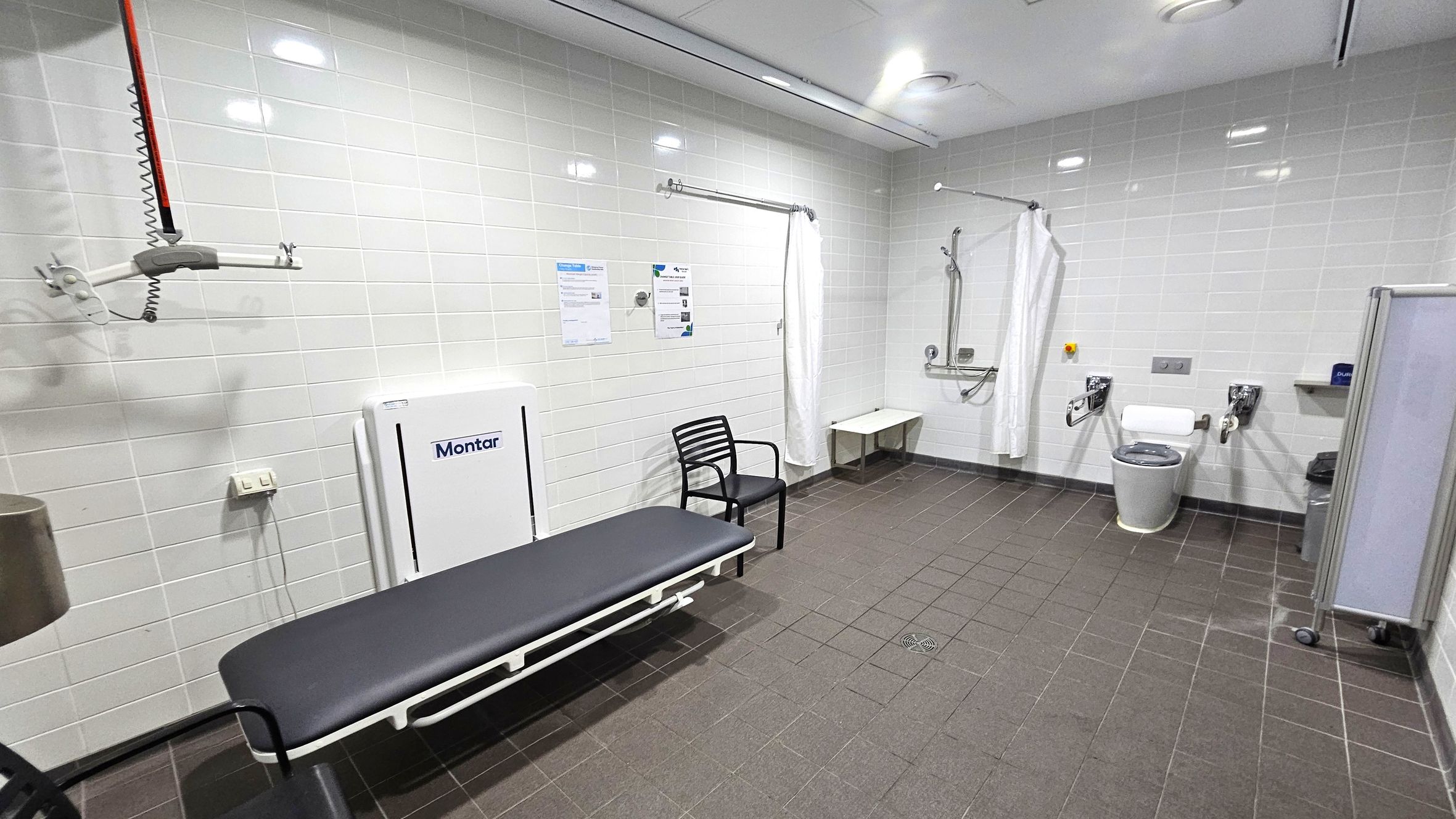 A spacious, accessible bathroom with white tiled walls and a dark tiled floor. On the left, there's a black bench with a white frame, a black chair, and a wall-mounted hoist system. In the centre, a white curtain hangs from a rail. On the right, there's a toilet with support bars, a small shelf, and a bin. The ceiling is white with recessed lighting. A foldable changing table is mounted on the wall. A spacious, accessible bathroom with white tiled walls and a dark tiled floor. On the left, there's a black bench with a white frame, a black chair, and a wall-mounted hoist system. In the centre, a white curtain hangs from a rail. On the right, there's a toilet with support bars, a small shelf, and a bin. The ceiling is white with recessed lighting. A foldable changing table is mounted on the wall.