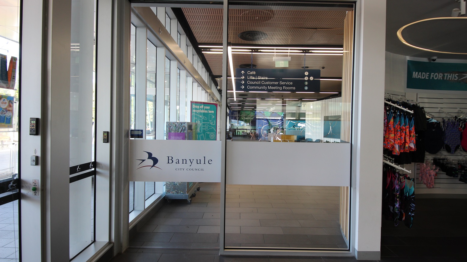 Glass entrance doors with a sign reading "Banyule City Council" in the centre. To the left, a keypad and glass panels. Inside, a directory sign points to a café, lifts, stairs, council customer service, and community meeting rooms. The interior has a wooden ceiling and visible lighting. On the right, a retail area displays colourful clothing on racks. Glass entrance doors with a sign reading "Banyule City Council" in the centre. To the left, a keypad and glass panels. Inside, a directory sign points to a café, lifts, stairs, council customer service, and community meeting rooms. The interior has a wooden ceiling and visible lighting. On the right, a retail area displays colourful clothing on racks.