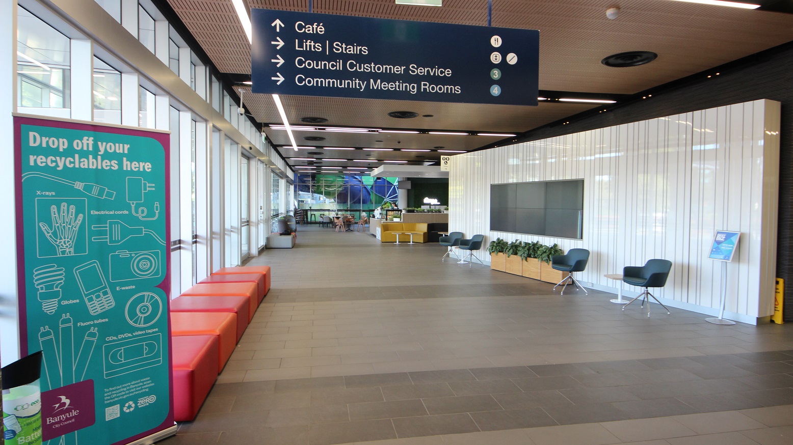 A spacious indoor corridor with a high ceiling featuring linear lights. On the left, large windows allow natural light to flood in, with a teal sign for recyclables and red seating along the wall. On the right, a white panelled wall with a mounted screen, flanked by two chairs and a row of potted plants. Overhead, a blue sign directs to a café, lifts, council customer service, and meeting rooms. In the background, a café area with tables and chairs is visible. A spacious indoor corridor with a high ceiling featuring linear lights. On the left, large windows allow natural light to flood in, with a teal sign for recyclables and red seating along the wall. On the right, a white panelled wall with a mounted screen, flanked by two chairs and a row of potted plants. Overhead, a blue sign directs to a café, lifts, council customer service, and meeting rooms. In the background, a café area with tables and chairs is visible.