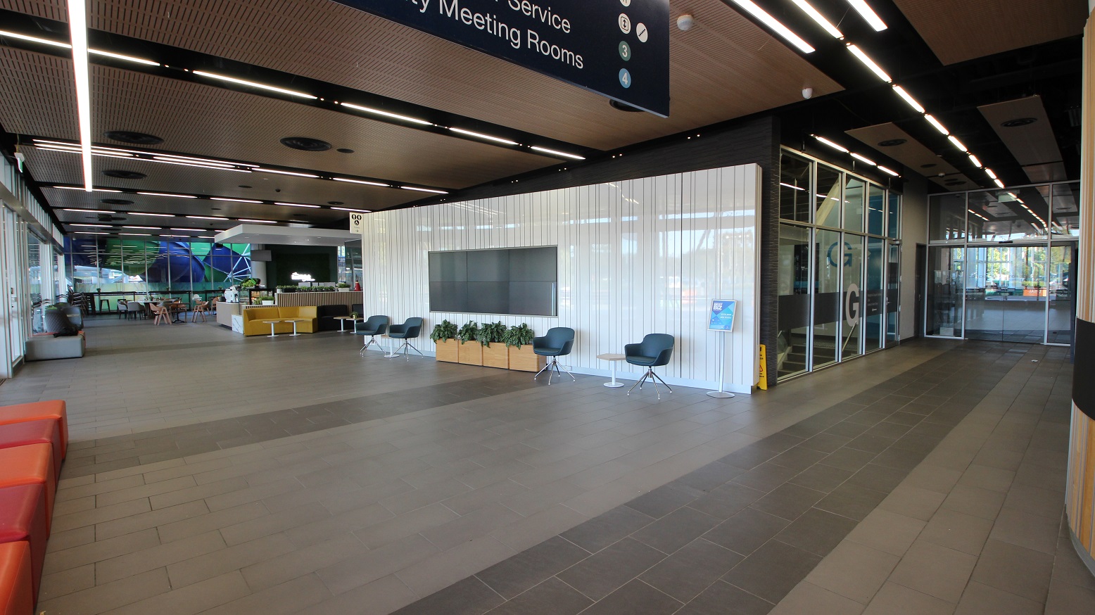 A modern interior space with a high ceiling featuring linear lights. On the right, there's a glass entrance with a sign above. In the centre, a white wall with a large screen is flanked by chairs and potted plants. On the left, a seating area with orange couches and a reception desk is visible. The floor is tiled in shades of grey, and the ceiling has a wood panel design with circular vents. A modern interior space with a high ceiling featuring linear lights. On the right, there's a glass entrance with a sign above. In the centre, a white wall with a large screen is flanked by chairs and potted plants. On the left, a seating area with orange couches and a reception desk is visible. The floor is tiled in shades of grey, and the ceiling has a wood panel design with circular vents.