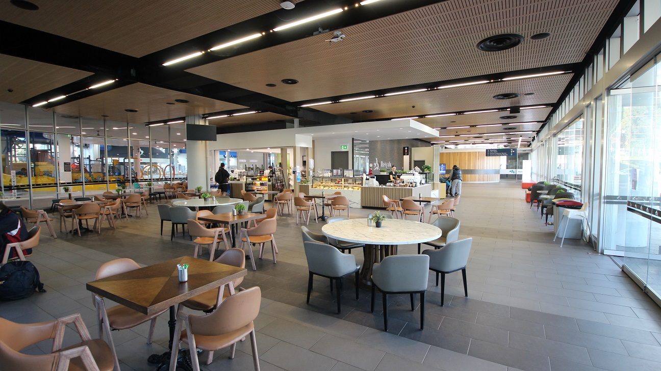 A spacious, modern café area with a variety of seating options, including round and square tables surrounded by wooden chairs. The left side features a glass wall with a view of an escalator and some equipment. In the centre, there's a counter with food displays and a person standing nearby. The right side has large windows allowing natural light to fill the space, with additional seating along the window. The ceiling has a wooden finish with black beams and recessed lighting. A spacious, modern café area with a variety of seating options, including round and square tables surrounded by wooden chairs. The left side features a glass wall with a view of an escalator and some equipment. In the centre, there's a counter with food displays and a person standing nearby. The right side has large windows allowing natural light to fill the space, with additional seating along the window. The ceiling has a wooden finish with black beams and recessed lighting.