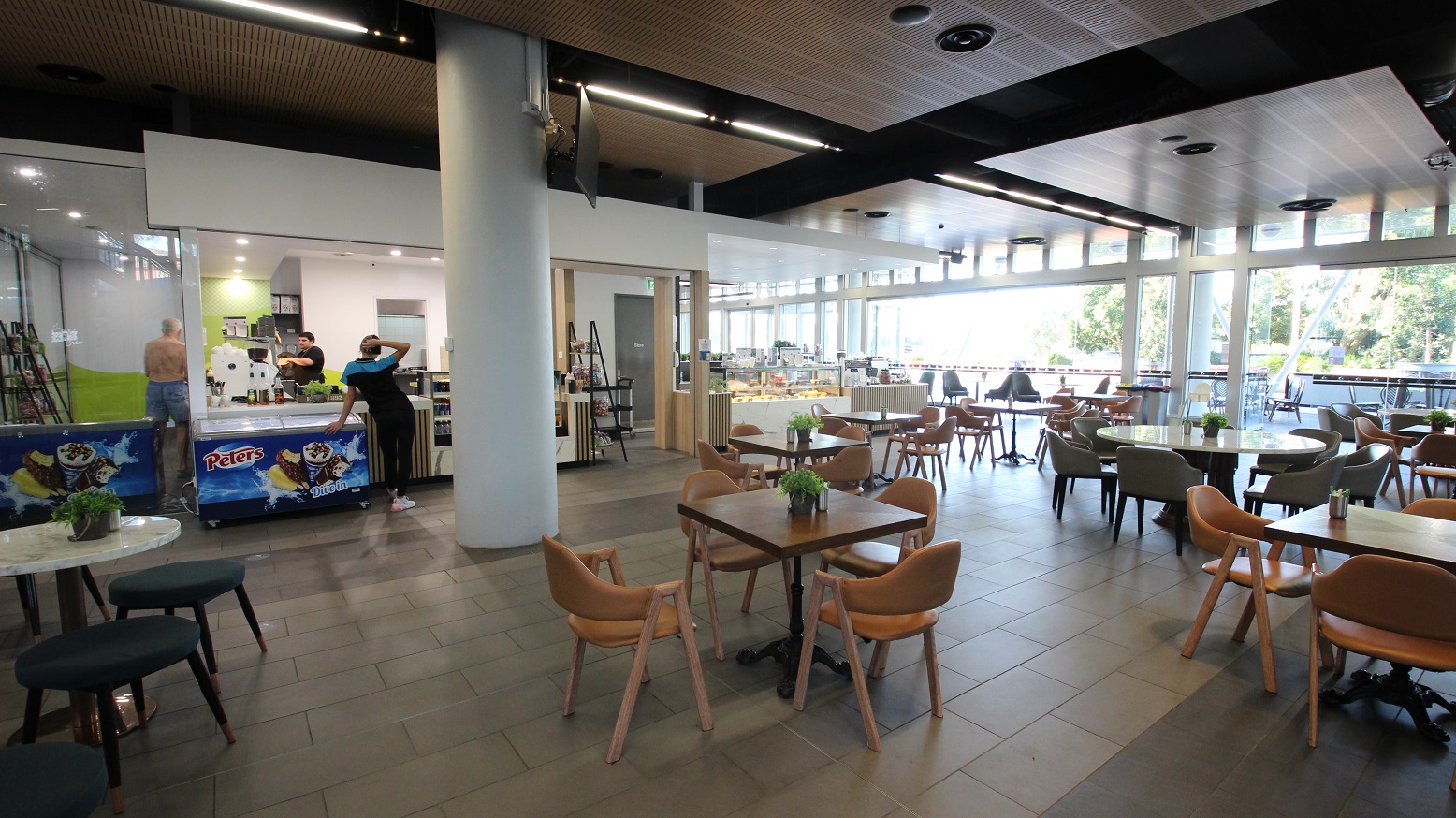 A modern café interior with a spacious seating area. On the left, there is a counter with ice cream freezers branded "Peters" and staff behind the counter. In the middle, several wooden tables and chairs are arranged neatly, each with a small plant as a centrepiece. The right side features large windows allowing natural light to flood in, with more seating visible outside. The ceiling has a mix of wooden and black panels, and the floor is tiled. A modern café interior with a spacious seating area. On the left, there is a counter with ice cream freezers branded "Peters" and staff behind the counter. In the middle, several wooden tables and chairs are arranged neatly, each with a small plant as a centrepiece. The right side features large windows allowing natural light to flood in, with more seating visible outside. The ceiling has a mix of wooden and black panels, and the floor is tiled.