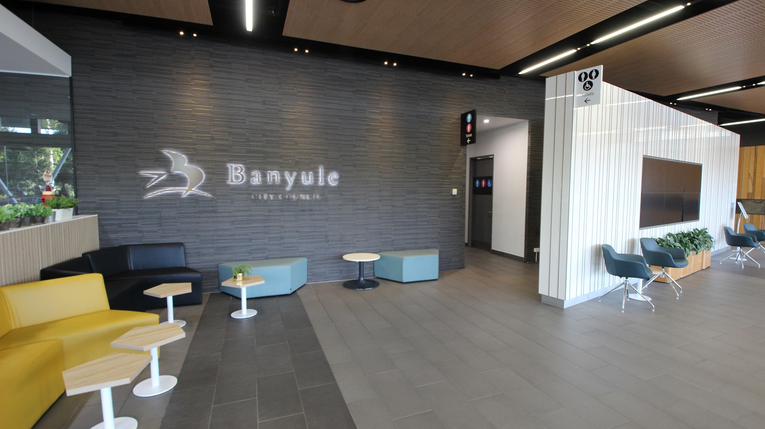 A modern lobby area with a grey tiled floor and a wooden panelled ceiling. On the left, there are yellow and black sofas with small hexagonal tables. The wall features the word "Banyule" with a bird logo. On the right, a white panelled wall has a mounted screen and blue chairs with a planter beneath. A corridor leads to restrooms, indicated by signs above. A window on the far left shows greenery outside. A modern lobby area with a grey tiled floor and a wooden panelled ceiling. On the left, there are yellow and black sofas with small hexagonal tables. The wall features the word "Banyule" with a bird logo. On the right, a white panelled wall has a mounted screen and blue chairs with a planter beneath. A corridor leads to restrooms, indicated by signs above. A window on the far left shows greenery outside.