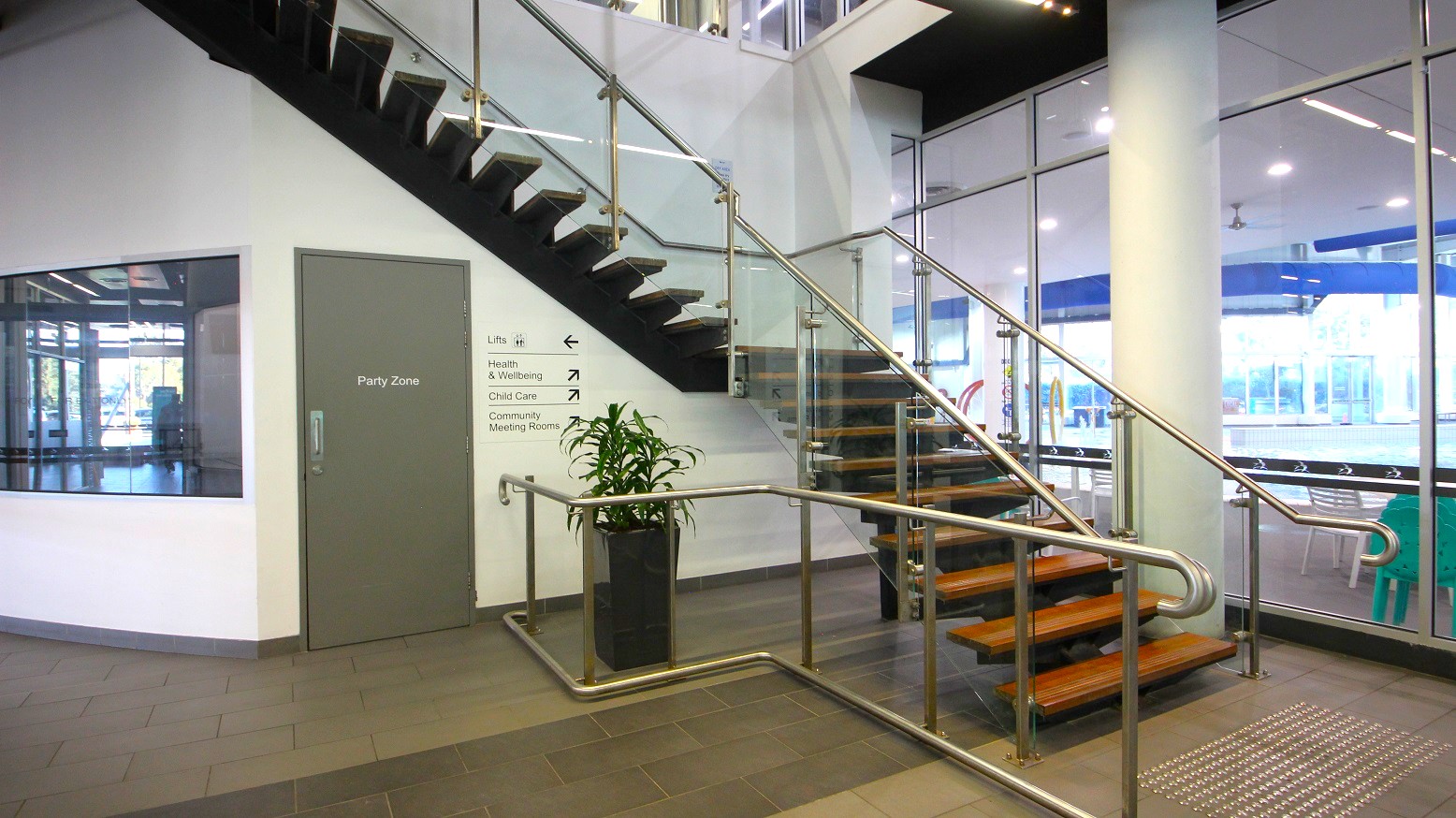 A modern indoor space with a staircase featuring wooden steps and glass railings. On the left, there's a grey door labelled "Party Zone" and a wall with directional signs. A potted plant sits near the base of the stairs. The floor is tiled, and large glass windows on the right reveal a view of an outdoor area with tables and chairs. The ceiling has recessed lighting. A modern indoor space with a staircase featuring wooden steps and glass railings. On the left, there's a grey door labelled "Party Zone" and a wall with directional signs. A potted plant sits near the base of the stairs. The floor is tiled, and large glass windows on the right reveal a view of an outdoor area with tables and chairs. The ceiling has recessed lighting.